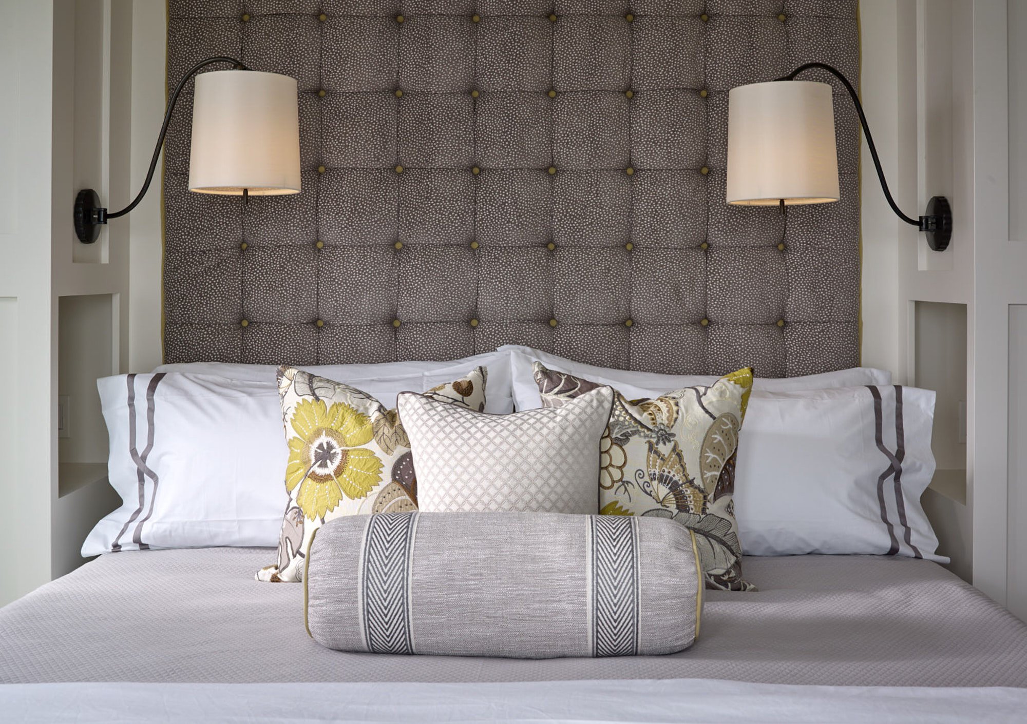 View of a neatly made bed with decorative pillows in front of a tufted headboard, with wall-mounted lamps on either side.