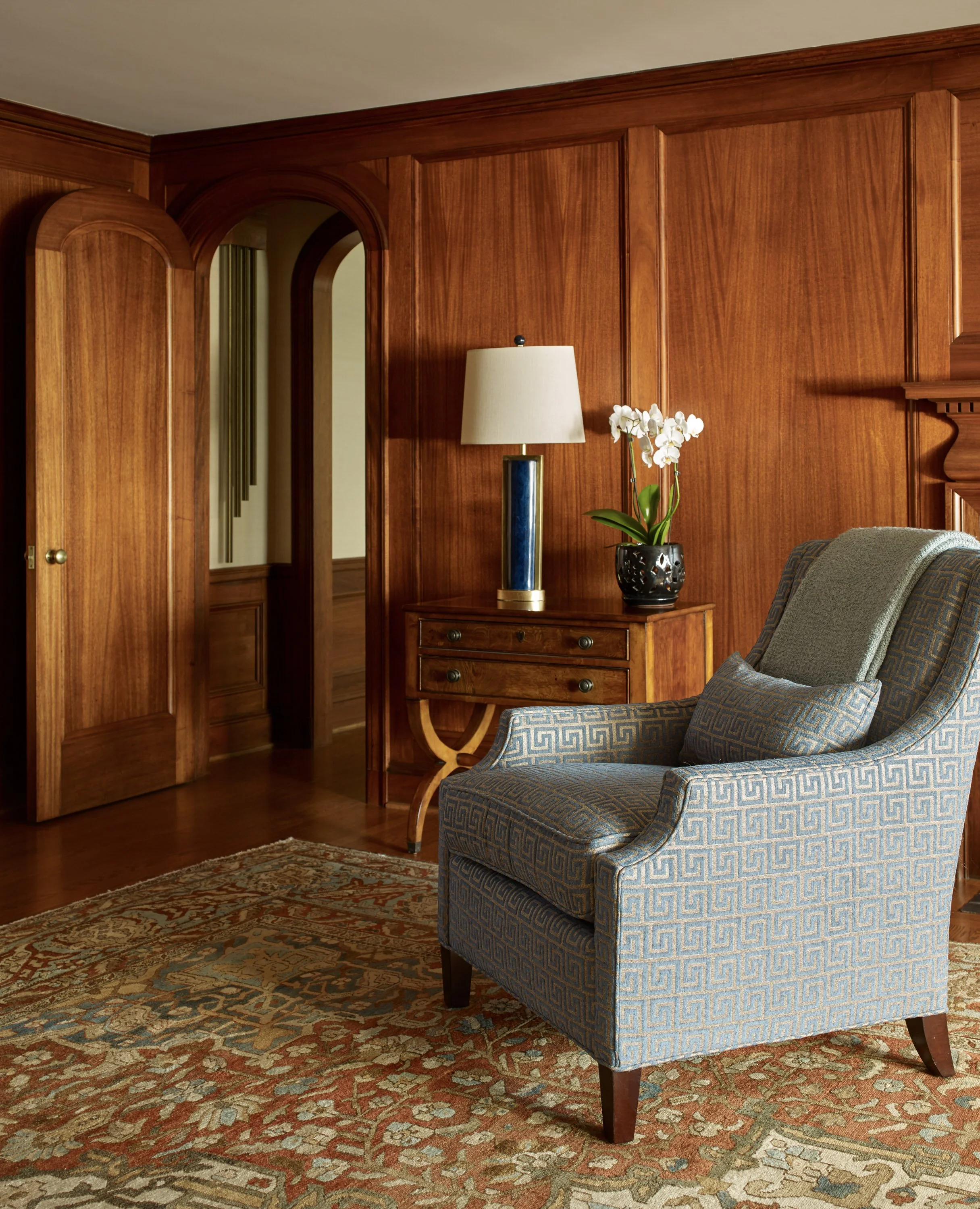 Elegant wood paneled living room with arched doorway, Asian rug in detailed warm tones, and contemporary club chair with light blue upholstery in geometric pattern.