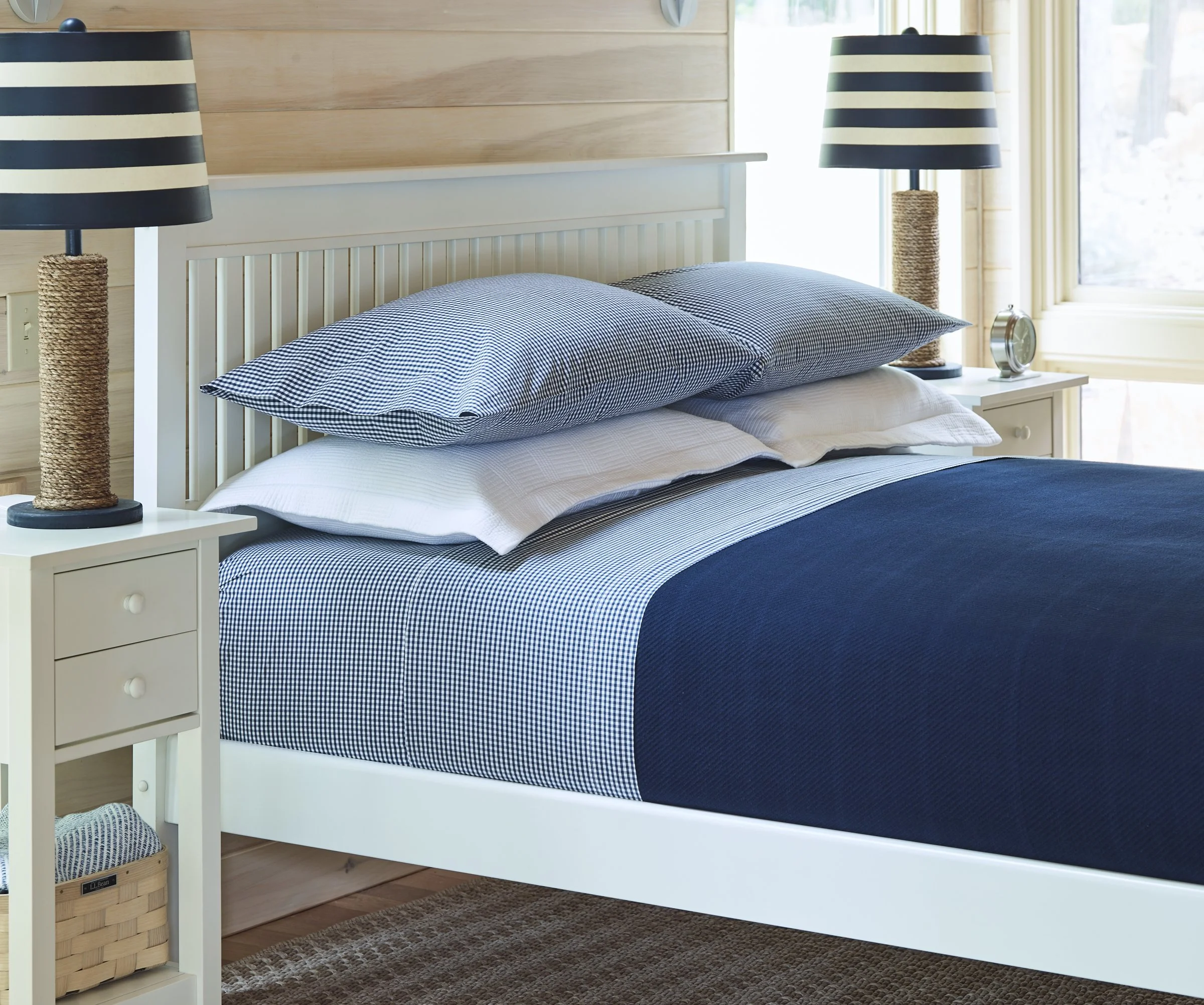 Close-up of a neatly made bed with striped navy and white bedding, flanked by matching striped table lamps on white nightstands, in a sunlit bedroom with wooden panel walls and a window in the background.