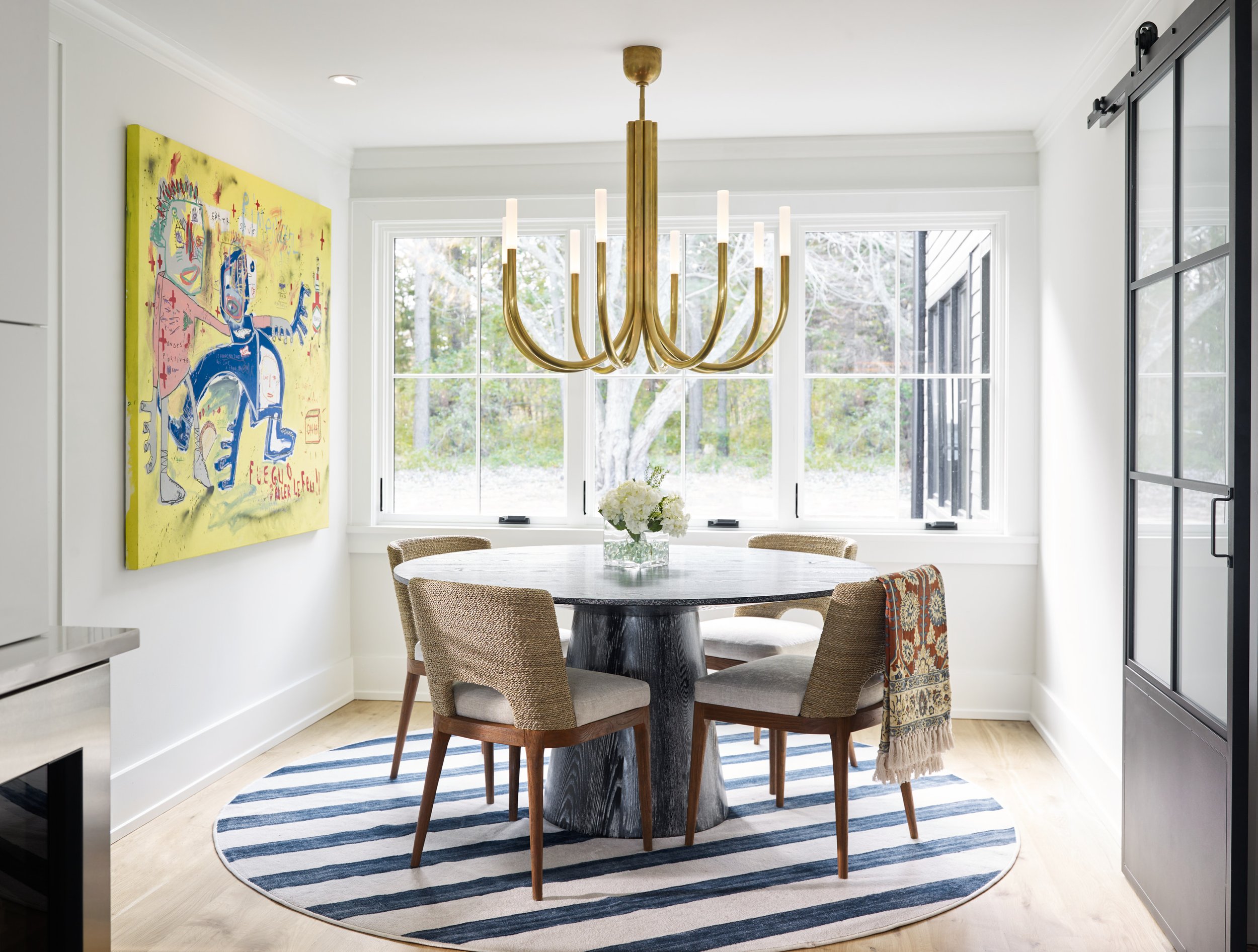 A dining area with a round, black, tree-trunk style table, surrounded by four woven armchairs, inside a room with large windows showing outdoor trees. A modern gold chandelier hangs over the table, and a colorful abstract artwork is on the wall to th