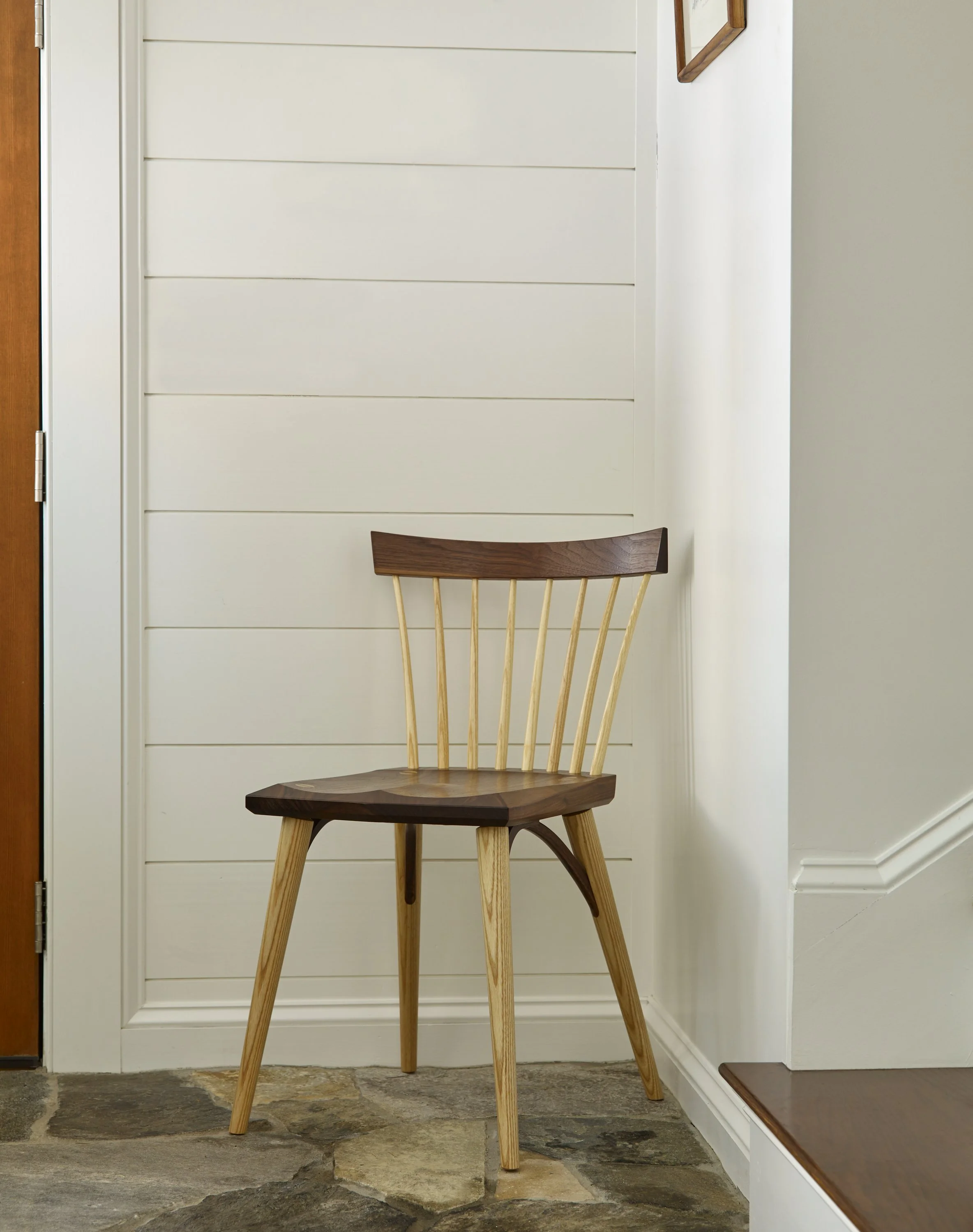 A wooden chair with a curved backrest and spindles, placed against a white paneled wall in a cozy hallway with stone flooring.