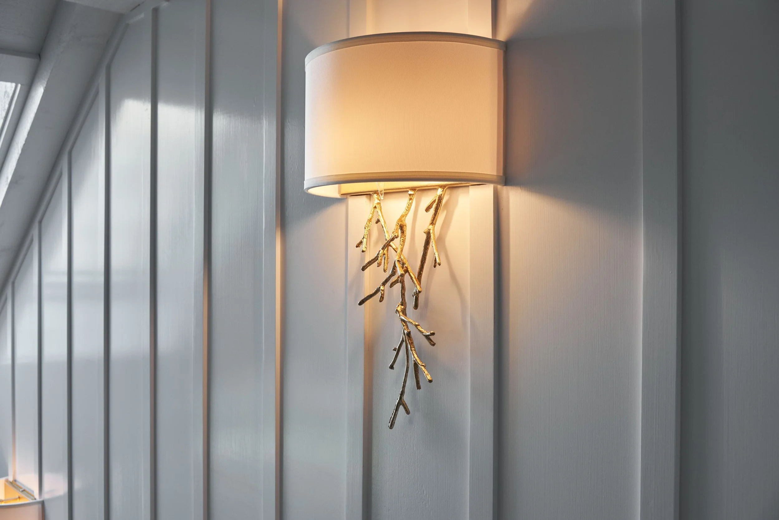 Wall-mounted lamp with a beige fabric shade and decorative gold branches on a gray wall.