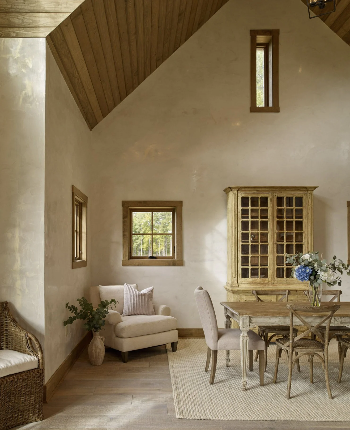 High ceiling pitched roof interior with rustic French country style dining table and chairs in warm tones, berber rug, and light cream walls covered with gold-veined Venetian plaster.
