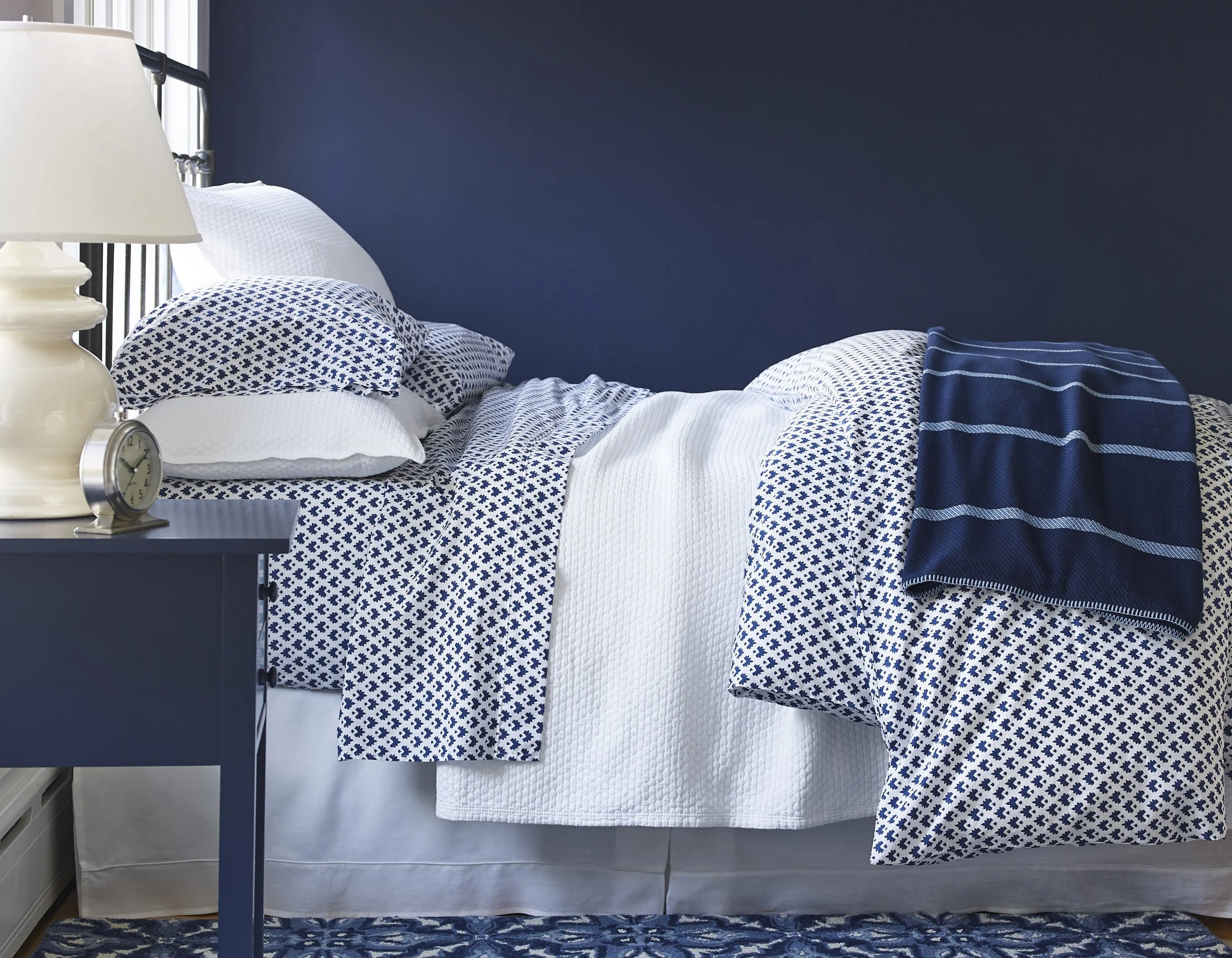 A bedroom with a bed covered in white and blue patterned linens, a dark blue wall, a white nightstand with a white lamp, a small clock, and a blue and white striped blanket.