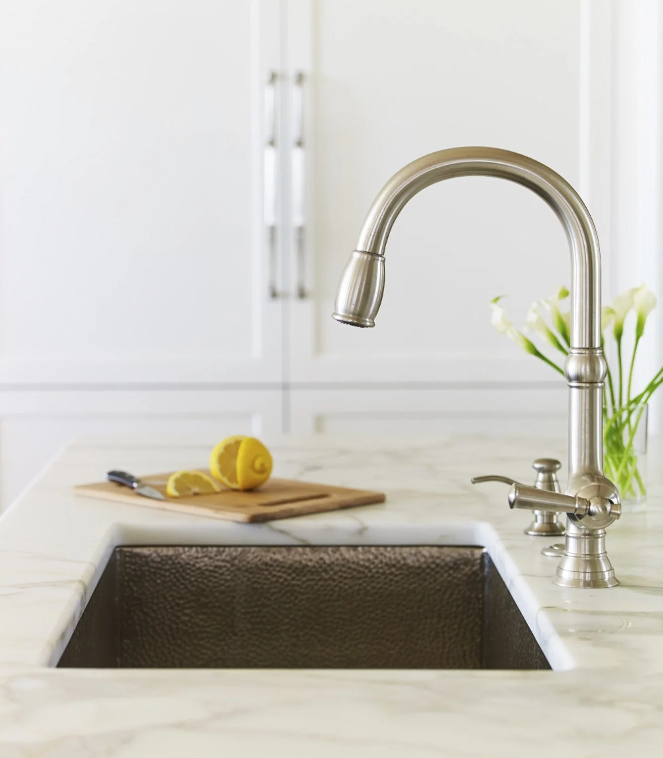 Close-up of a kitchen sink with a stainless steel faucet, a cutting board with lemon slices, and a vase with white flowers in the background.