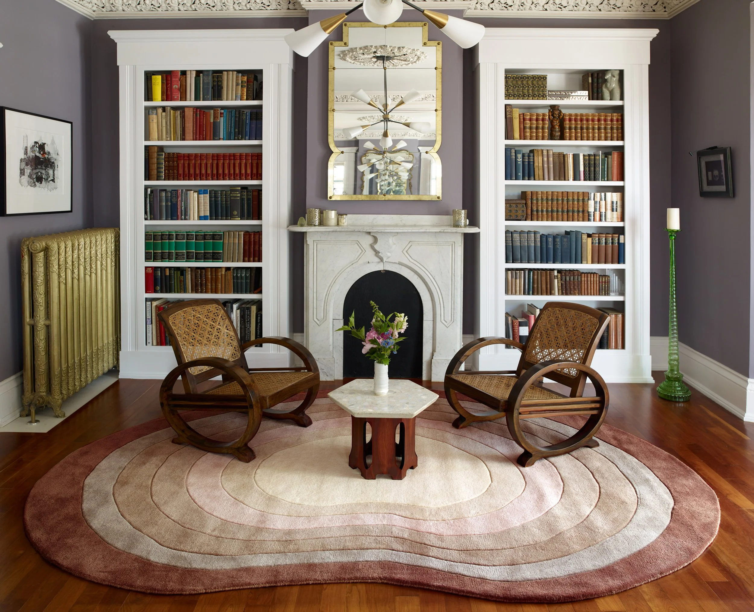A living room with grey walls, white built-in bookshelves filled with books, a white marble fireplace with a decorative mirror above it, and a round rug with pink, beige, and brown tones. There are four wooden chairs with woven cane seats around a sm