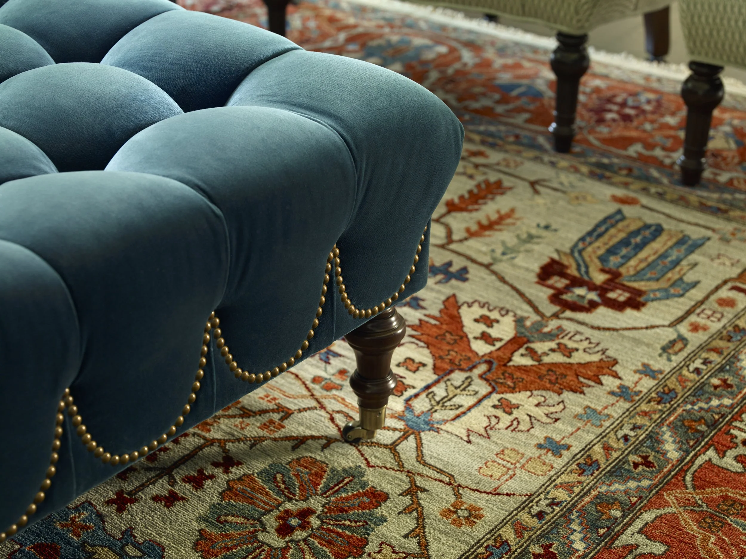 Close-up of a blue, plush, tufted armchair with brass nailhead trim, placed next to a traditional patterned area rug with orange, blue, beige, and red hues.
