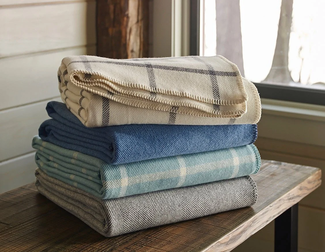 A stack of neatly folded plaid and solid-colored blankets on a wooden table next to a window.