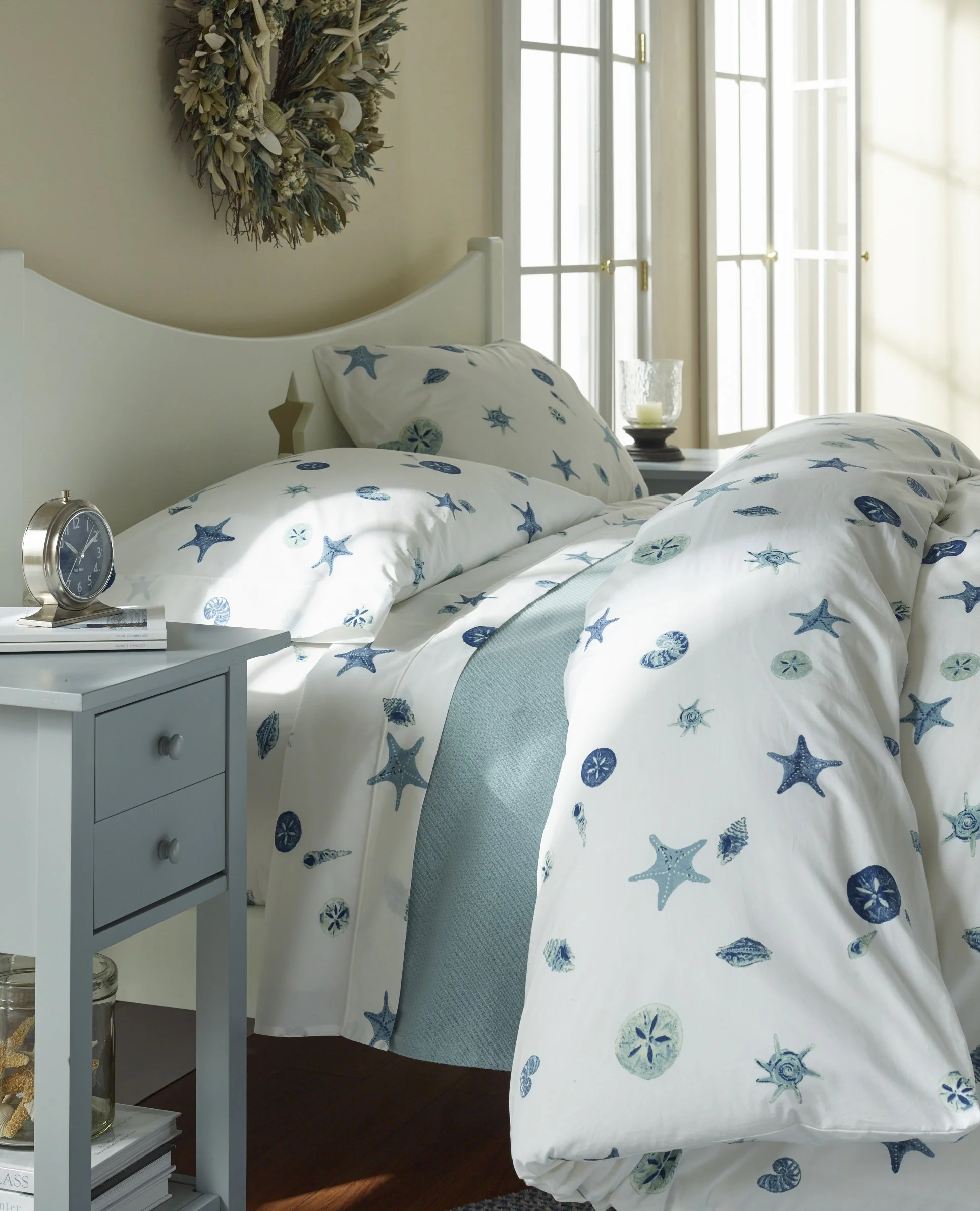 A cozy bedroom with a white bed covered with seashell and starfish patterned bedding, a bedside table with a clock and a candle in a glass holder, a large window with white framed glass panes letting in natural light, decorative dried flower arrangem