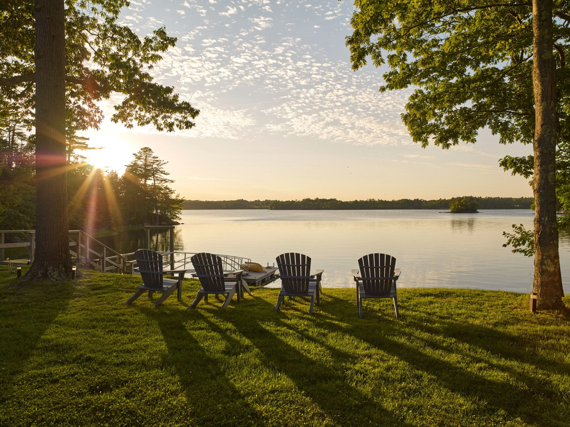 A lakeside scene during sunset with a few Adirondack chairs on a grassy area, trees framing the view, and the sun reflecting on the water.