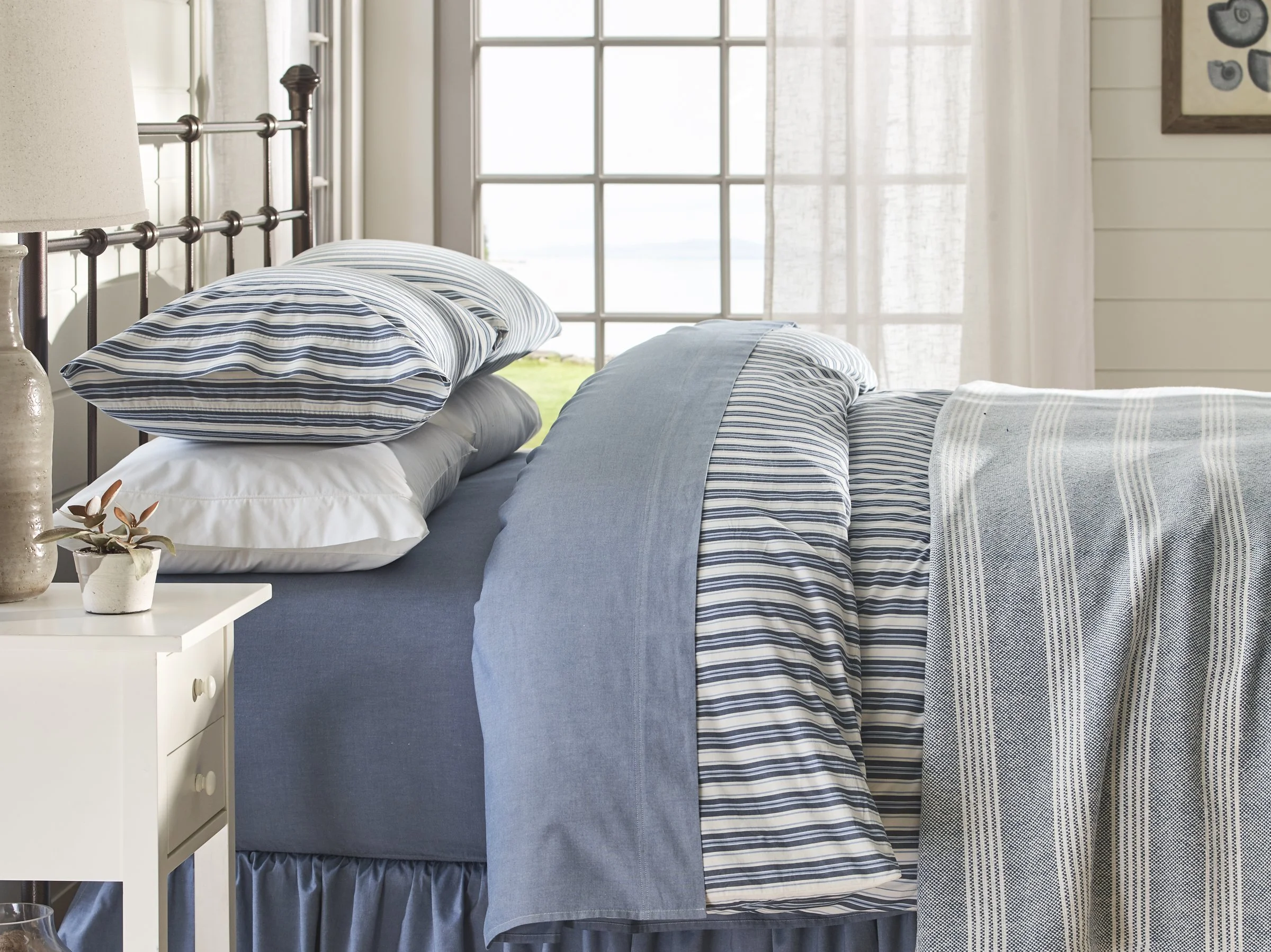 A neatly made bed with blue and white striped pillows and bedspread in a bright bedroom with large windows and white walls.