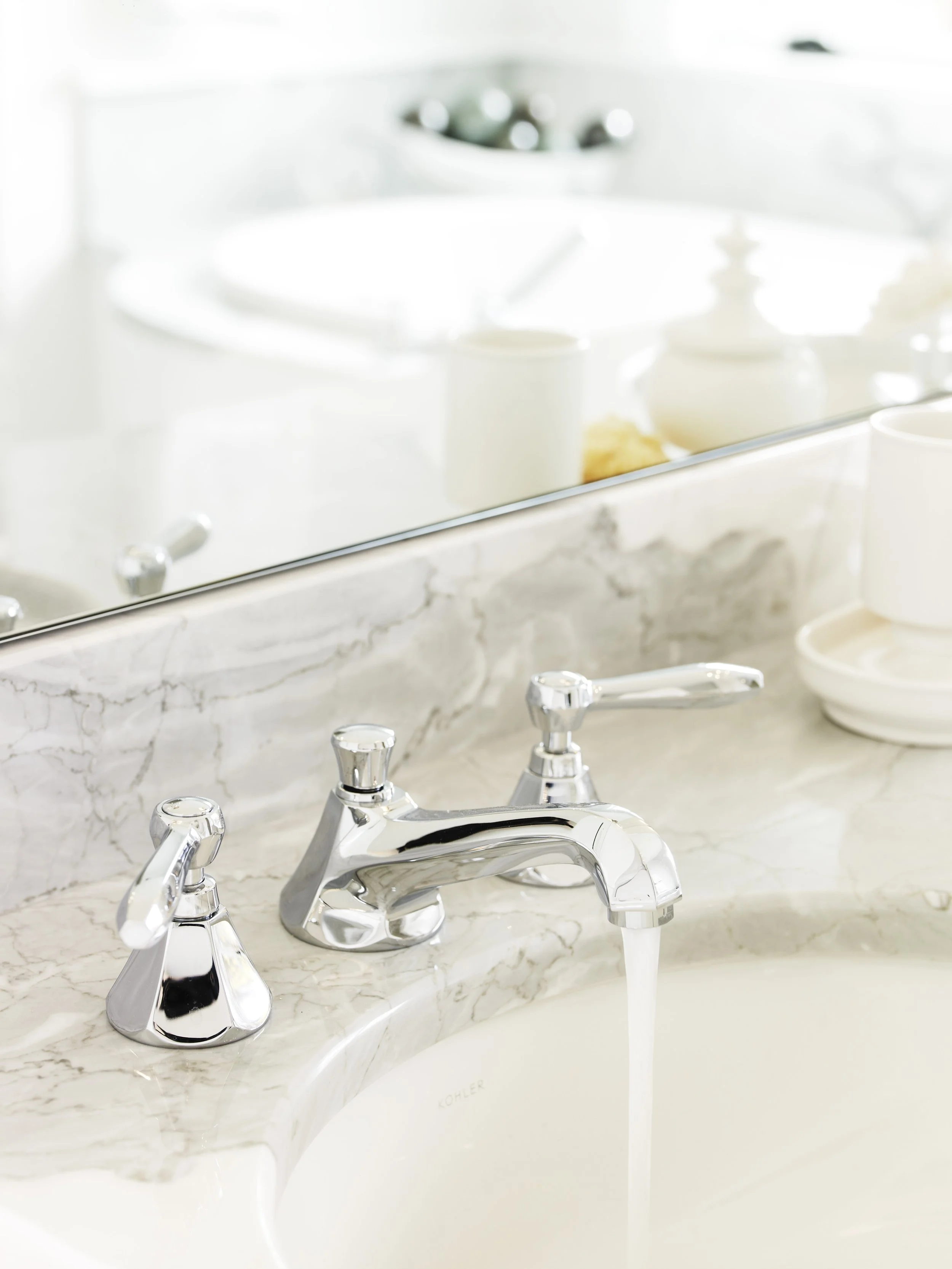 Close-up of a bathroom sink with a chrome faucet, marble countertop, and a mirror reflects objects including a soap dispenser and small decorative items.