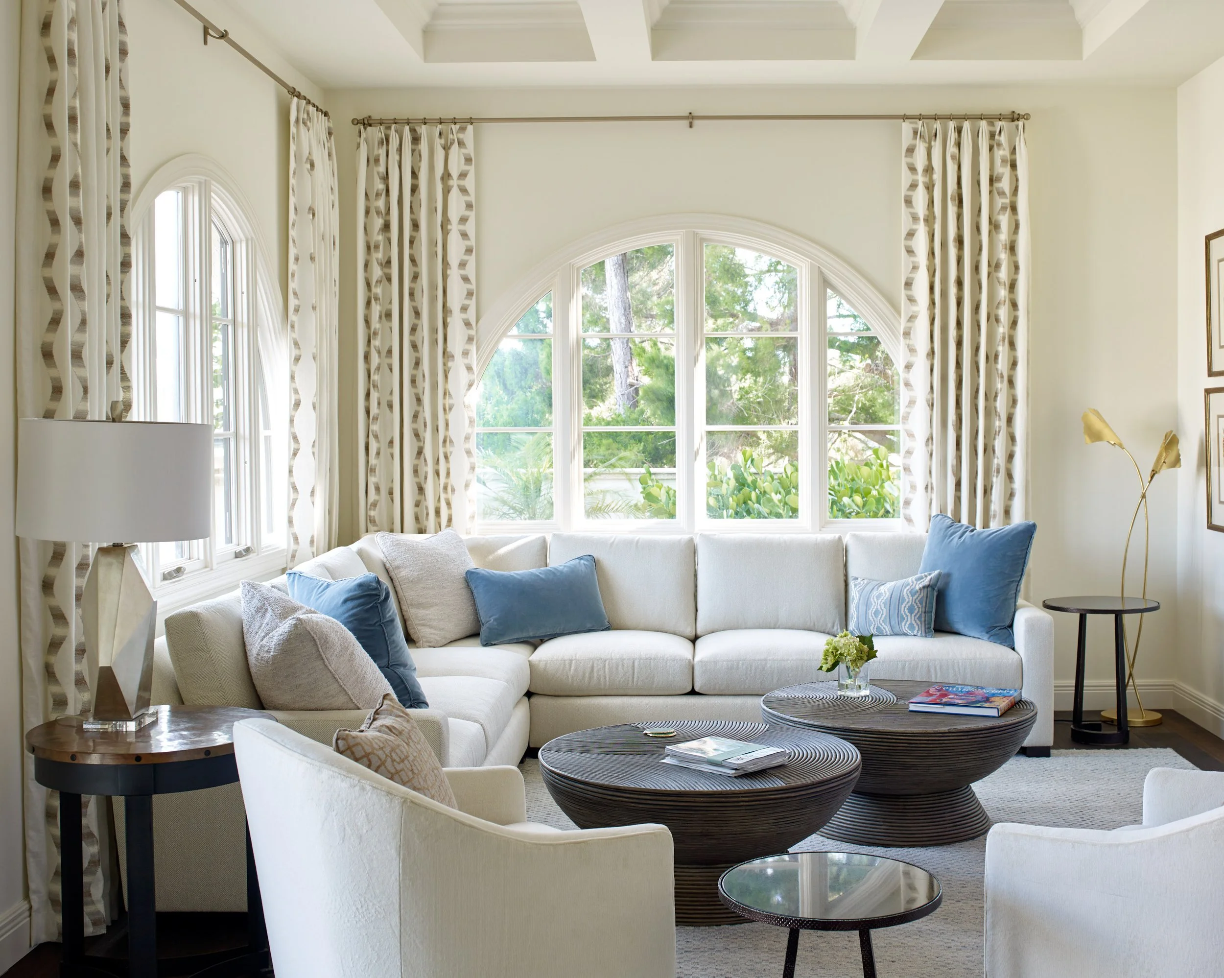 Living room with a large, curved window with white trim, beige curtains with a geometric pattern, white sofas with blue and beige pillows, a round wooden coffee table, a floor lamp with gold accents, and greenery visible outside.