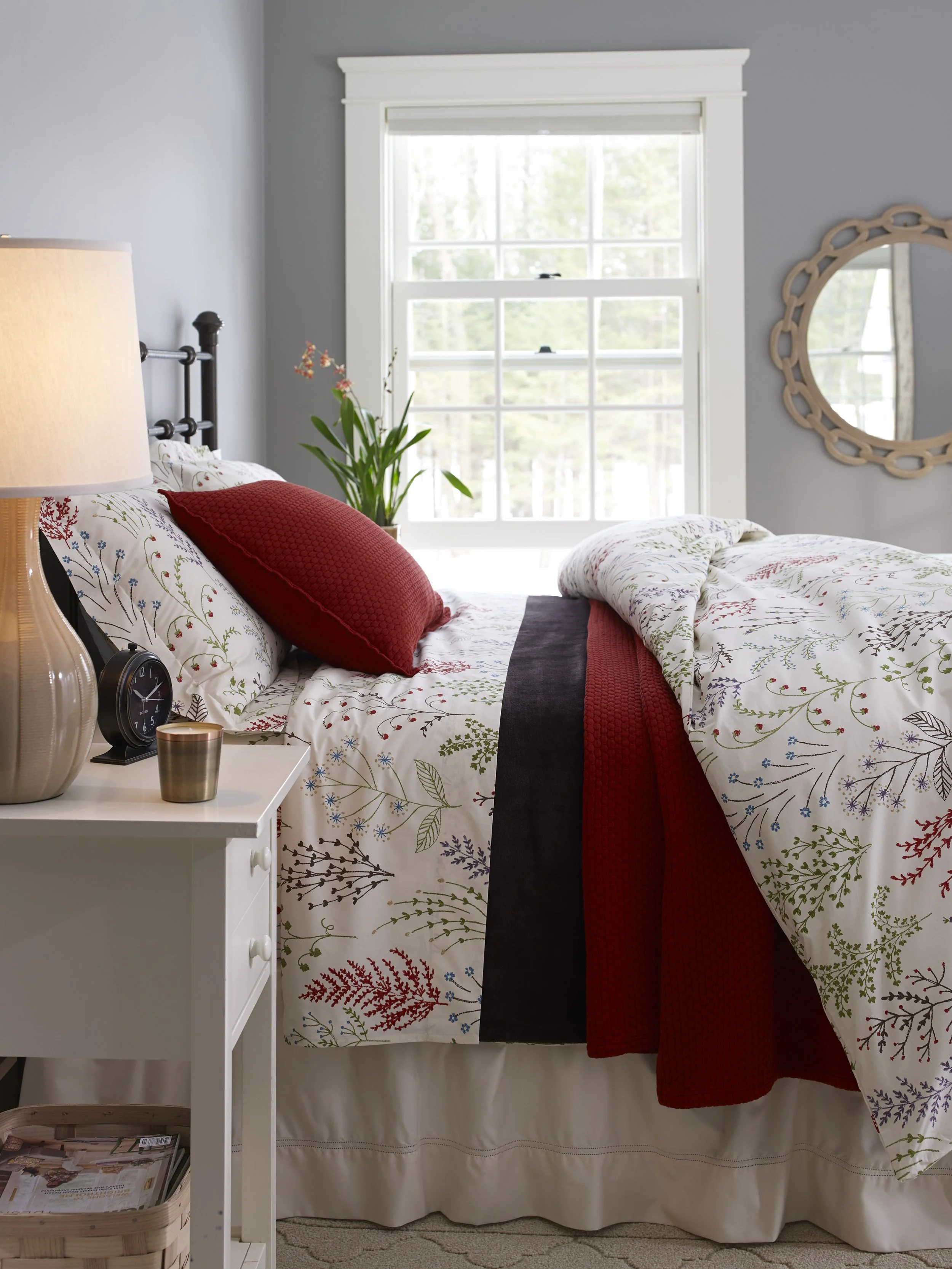 A cozy bedroom features a bed with floral-patterned bedding, a red pillow, and a black and red blanket. There's a white nightstand with a clock, a candle, and a lamp. Next to the bed is a basket of magazines. A window with white trim lets in natural 