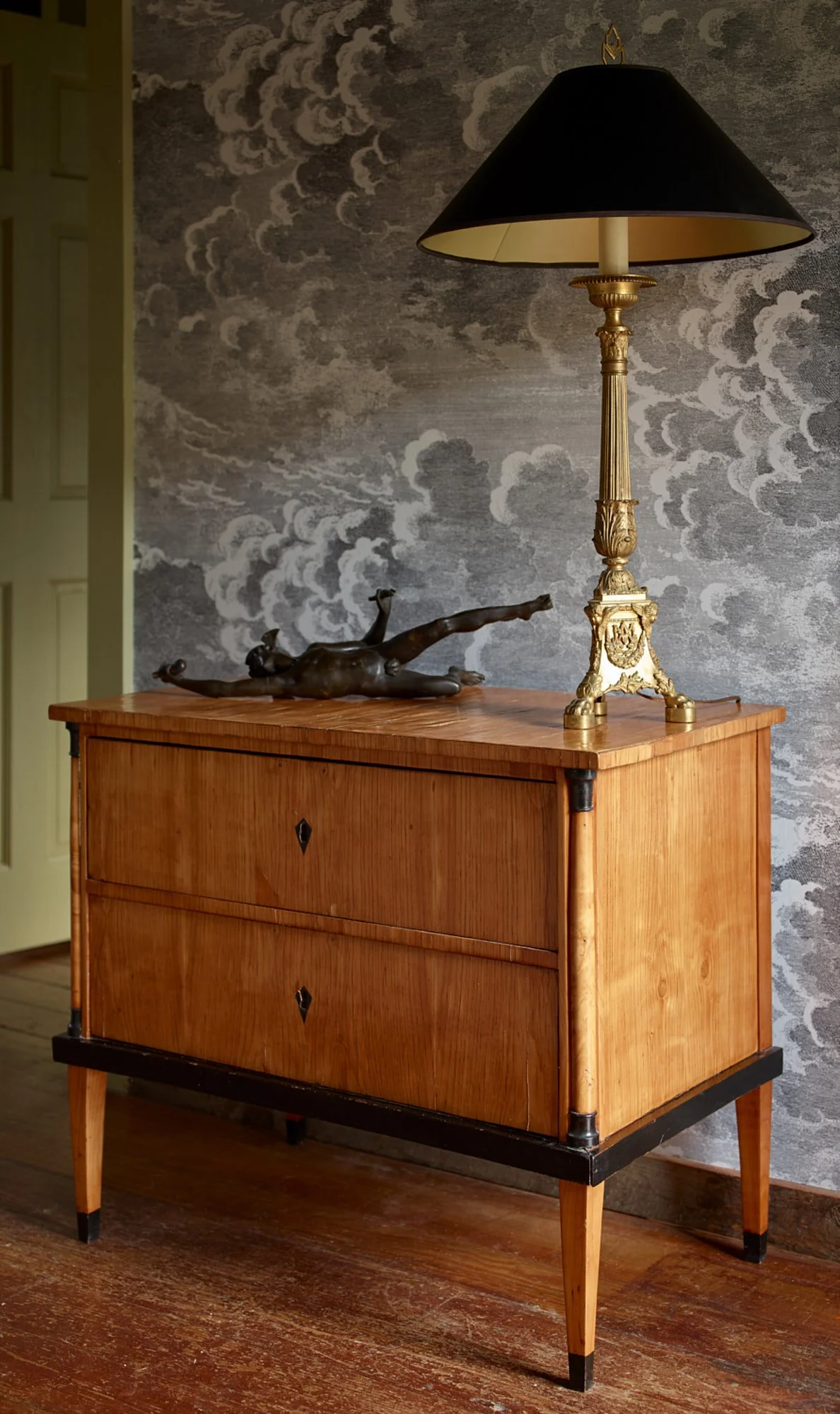 A wooden dresser with black and gold accents, a bronze sculpture of a reclining figure, and a tall ornate table lamp on top. The background features textured gray wallpaper with cloud patterns.