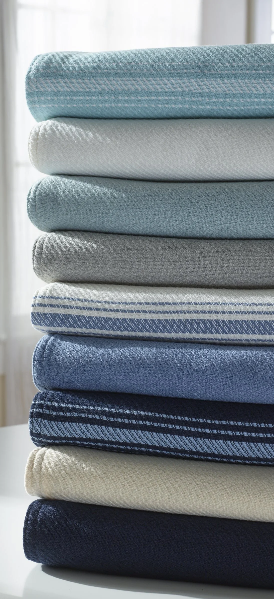 Stacked folded towels in various shades of blue, gray, and white with different patterns, placed on a white surface near a window.