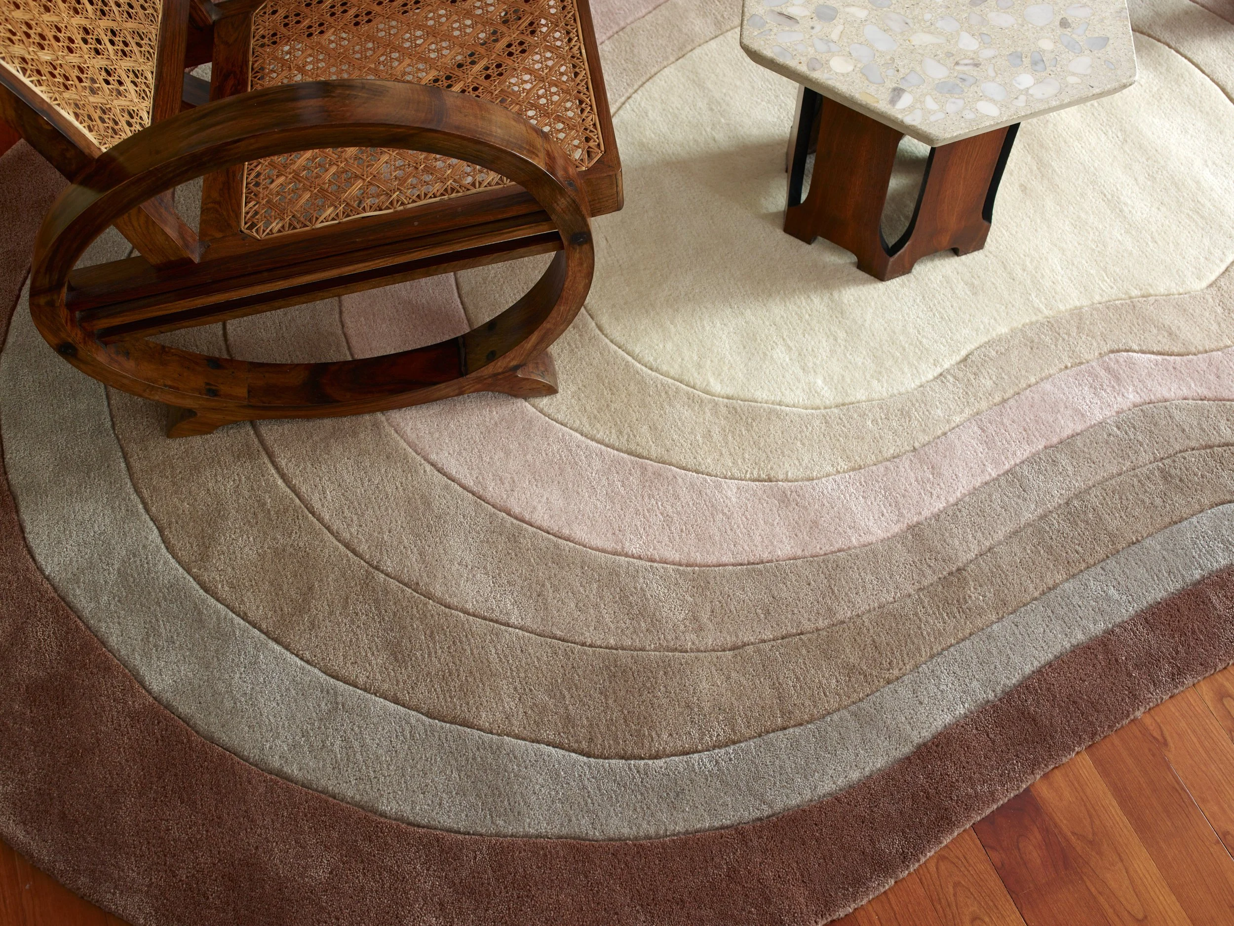 A beige and pink abstract patterned area rug with a surrounding wood floor, with two wooden chairs and a small square table with a terrazzo top.