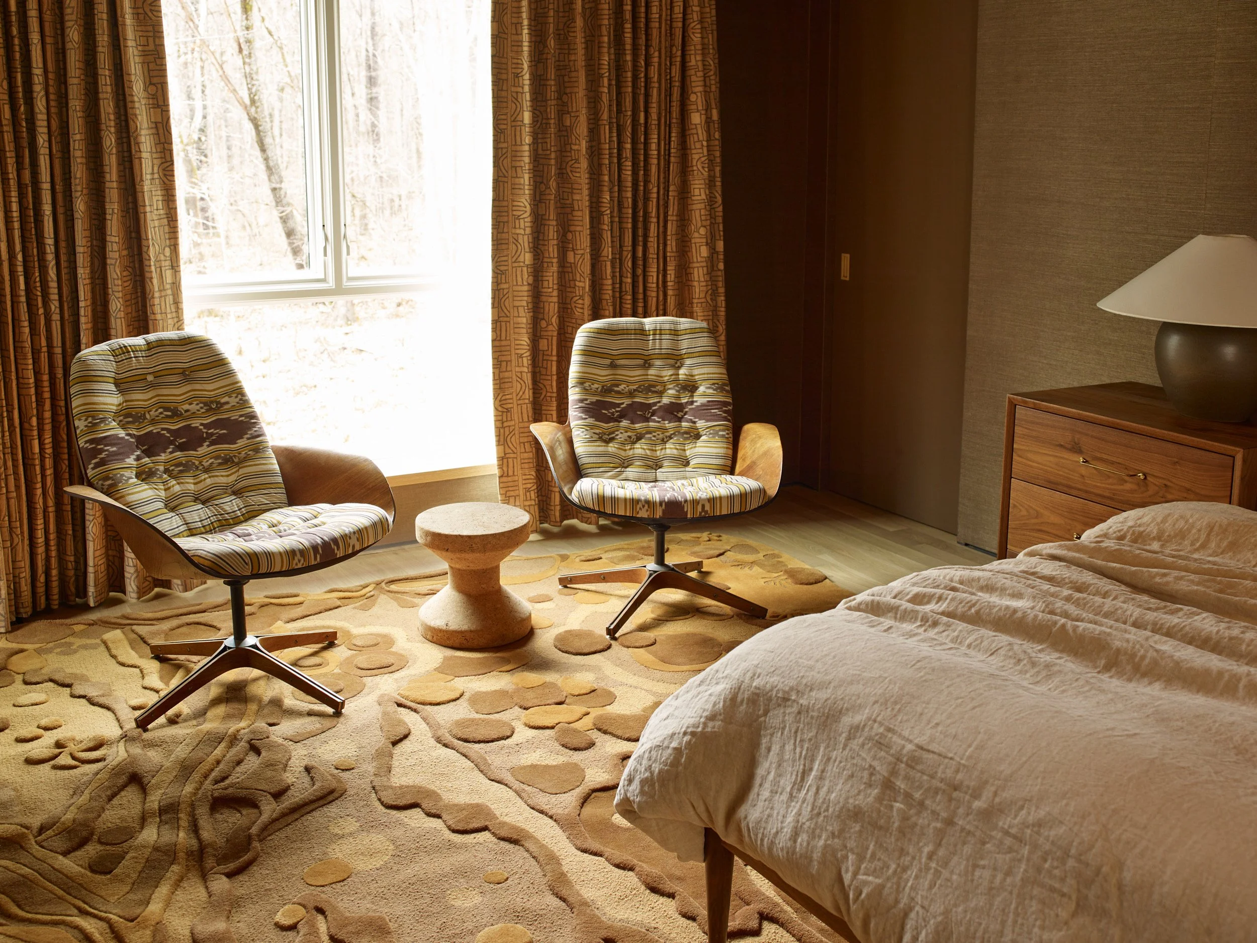 A hotel room with two striped armchairs, a small round table, a bed with beige sheets, a wooden dresser with a lamp, and a window with patterned curtains overlooking a wooded area.