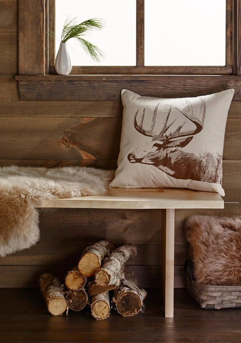 A cozy rustic indoor space with a wooden wall and window, a decorative cushion with a stag print, a stack of firewood, a sheepskin rug, a basket with faux fur, and a small vase with greenery on the windowsill.