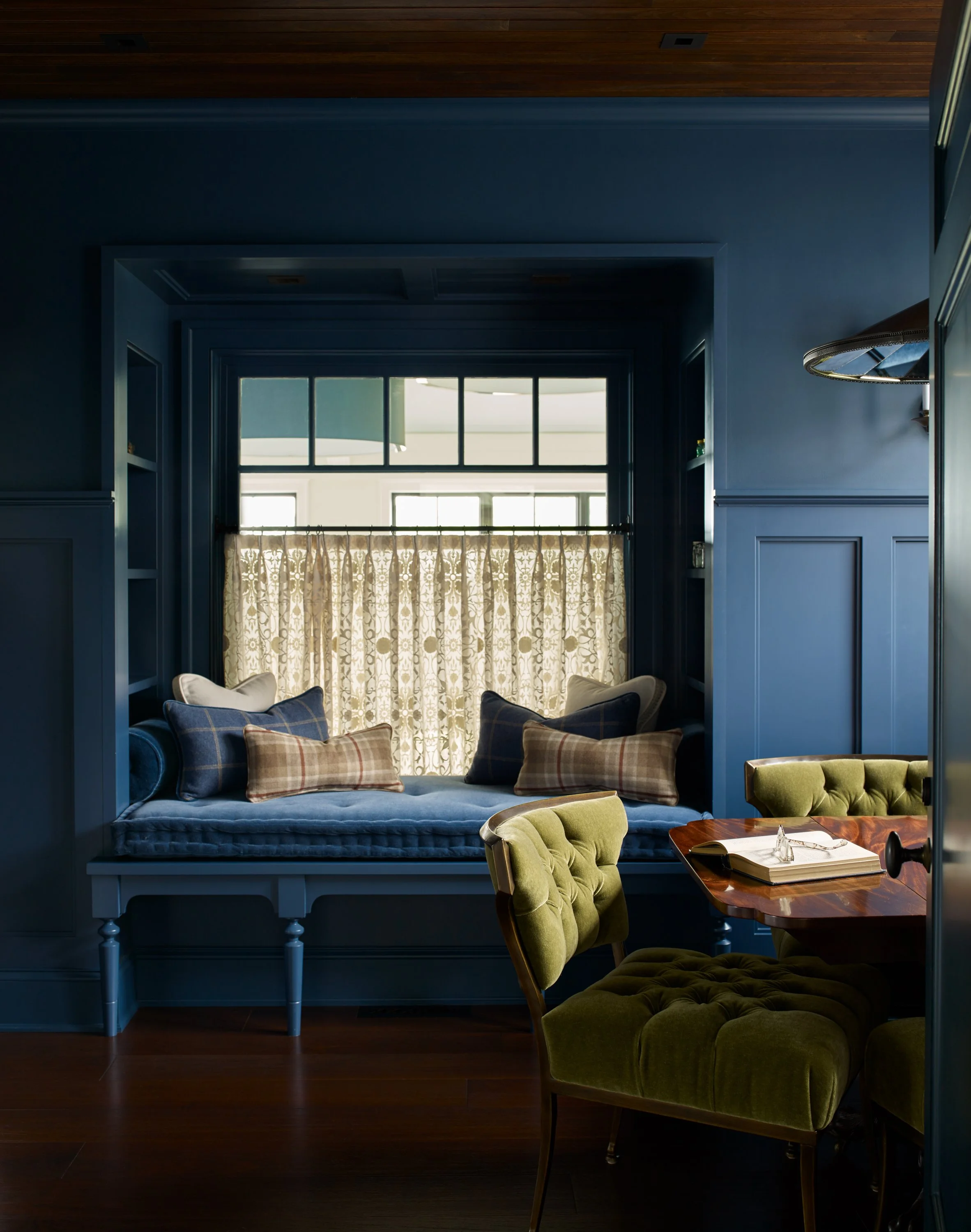 Cozy seating area with window seat enclosed by blue wood paneling, decorated with plaid pillows, and a window with curtains showing outdoor light, and a nearby wooden table with a green velvet armchair.
