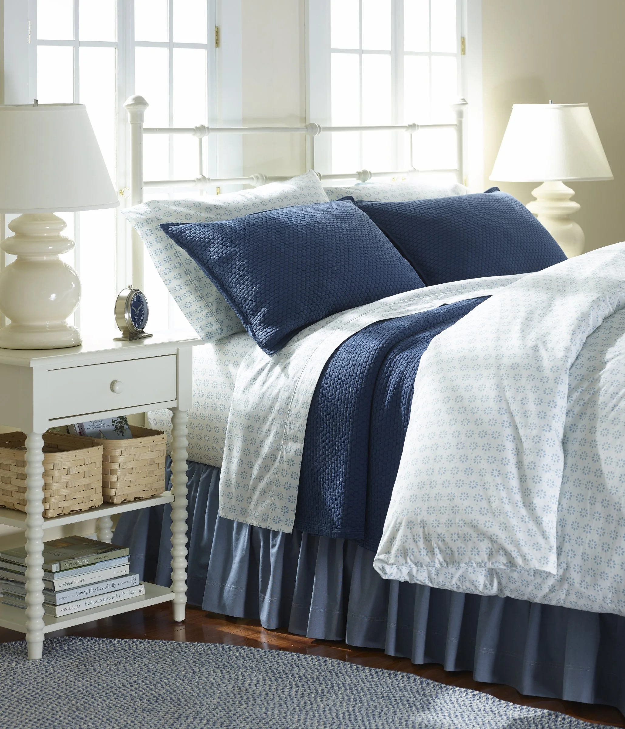 A bedroom with a white metal bed frame, blue and white bedding, and two table lamps on white nightstands. There are books on the lower shelf of the nightstand and a window with multiple panes behind the bed, letting in bright natural light. The floor