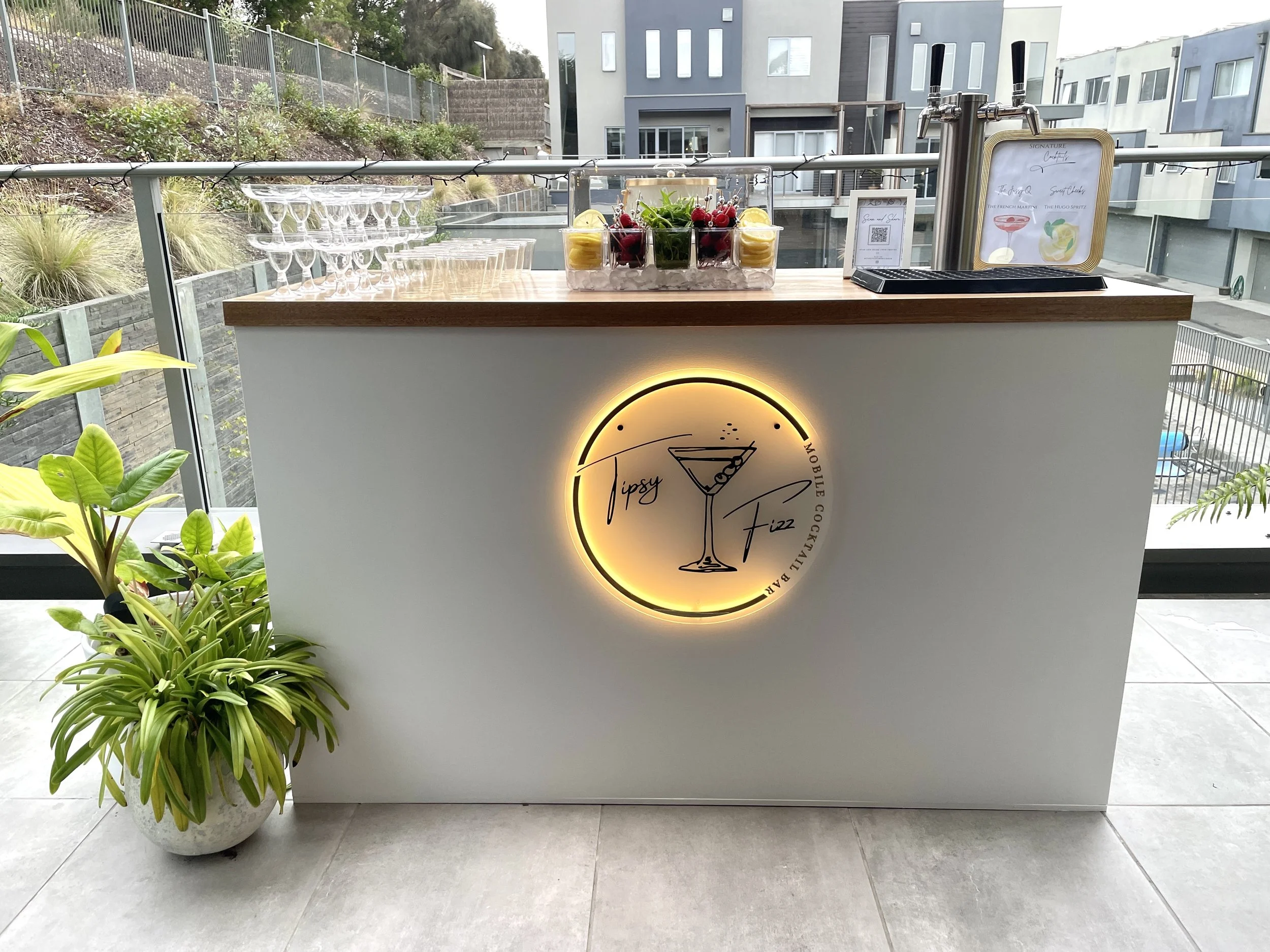 An outdoor bar setup on a balcony with a sign featuring a cocktail illustration, glassware, and a container of berries and lemon slices, with plants in the foreground.