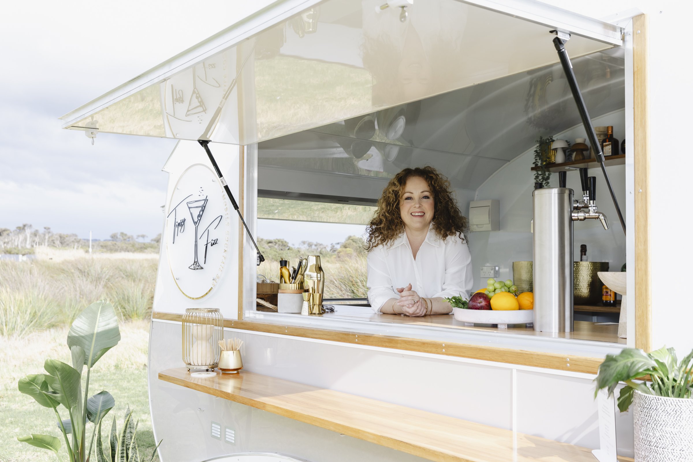 A smiling woman stands inside a mobile food truck with a wooden exterior and a large open window showing fresh fruits and bar tools, set against a rural outdoor background.