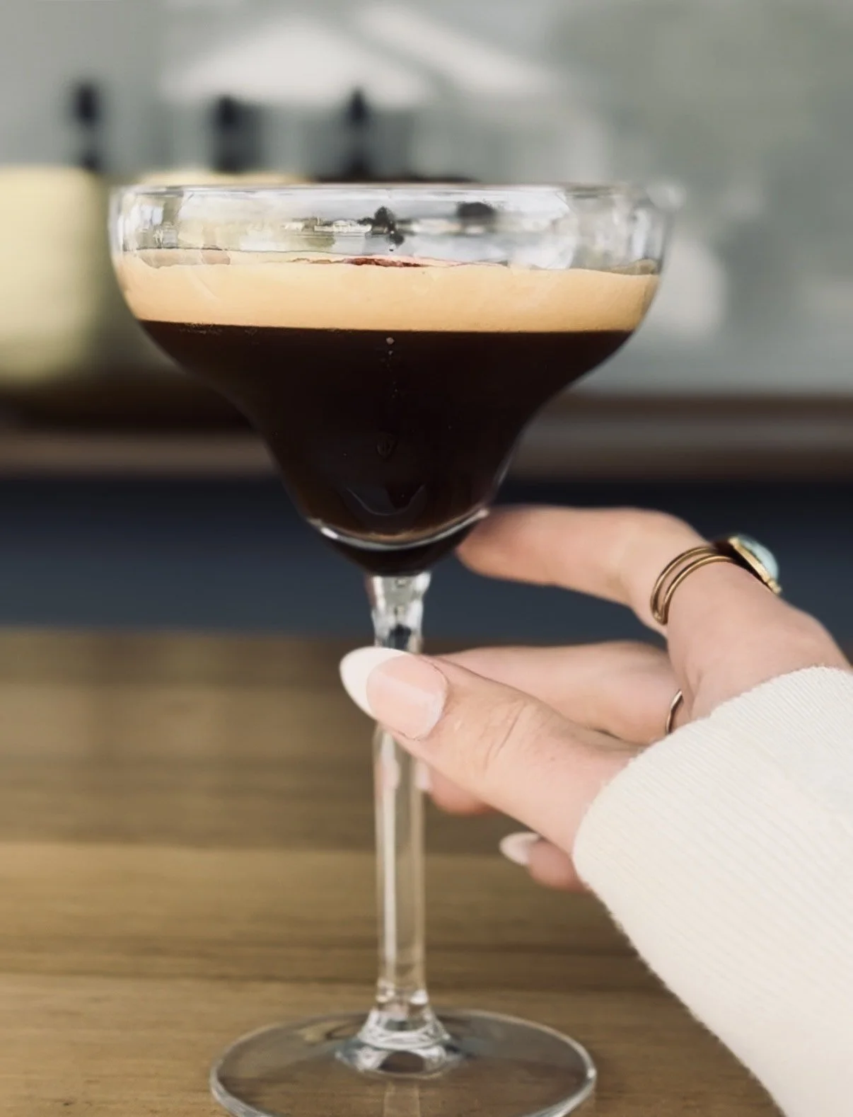 Close-up of a hand holding a martini glass with dark coffee-colored liquid, on a wooden surface background.