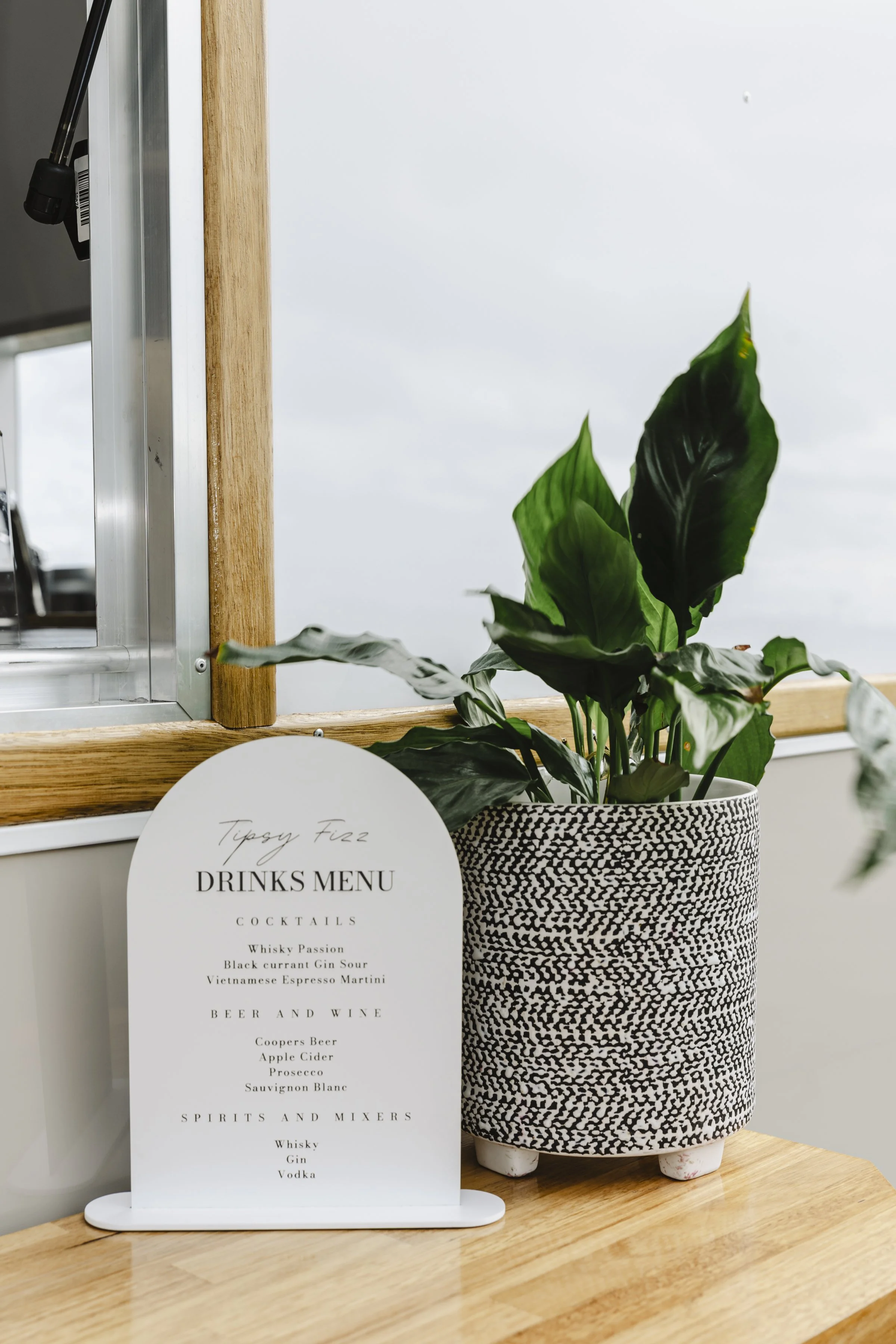 A drinks menu sign with cocktail options and a potted plant on a wooden table.