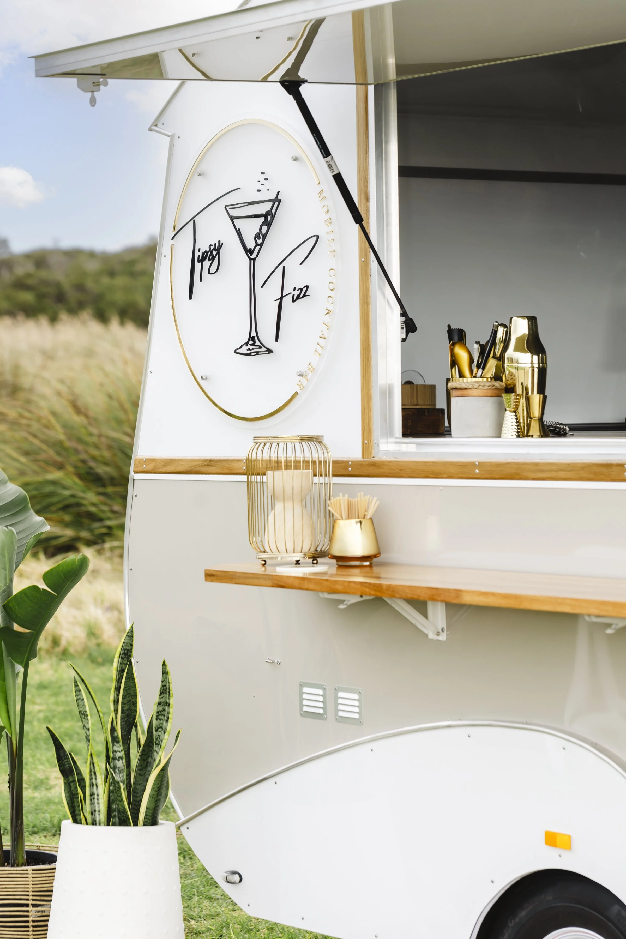 A white and wood food truck with a sign reading 'Tasty Fiz' and a drawing of a cocktail glass. The truck has a wooden shelf with a gold-colored container and a wire lantern. Potted plants are in front of the truck, with a grassy field and trees in the background.