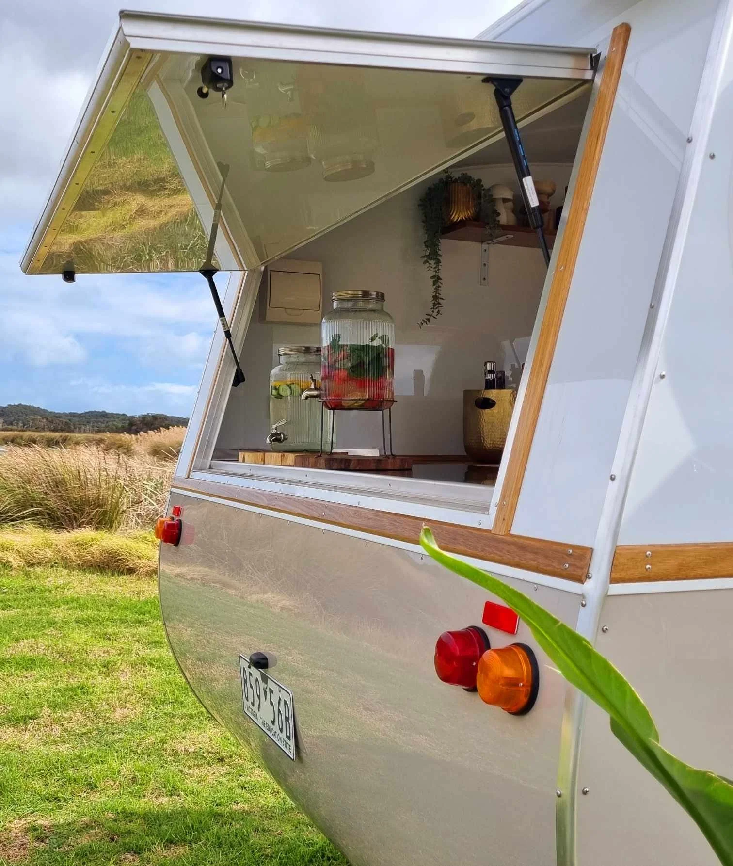 A compact mobile food trailer with an open window serving area, showing two glass beverage dispensers with fruit and herbs inside, set outdoors on a grassy area with a cloudy sky in the background.