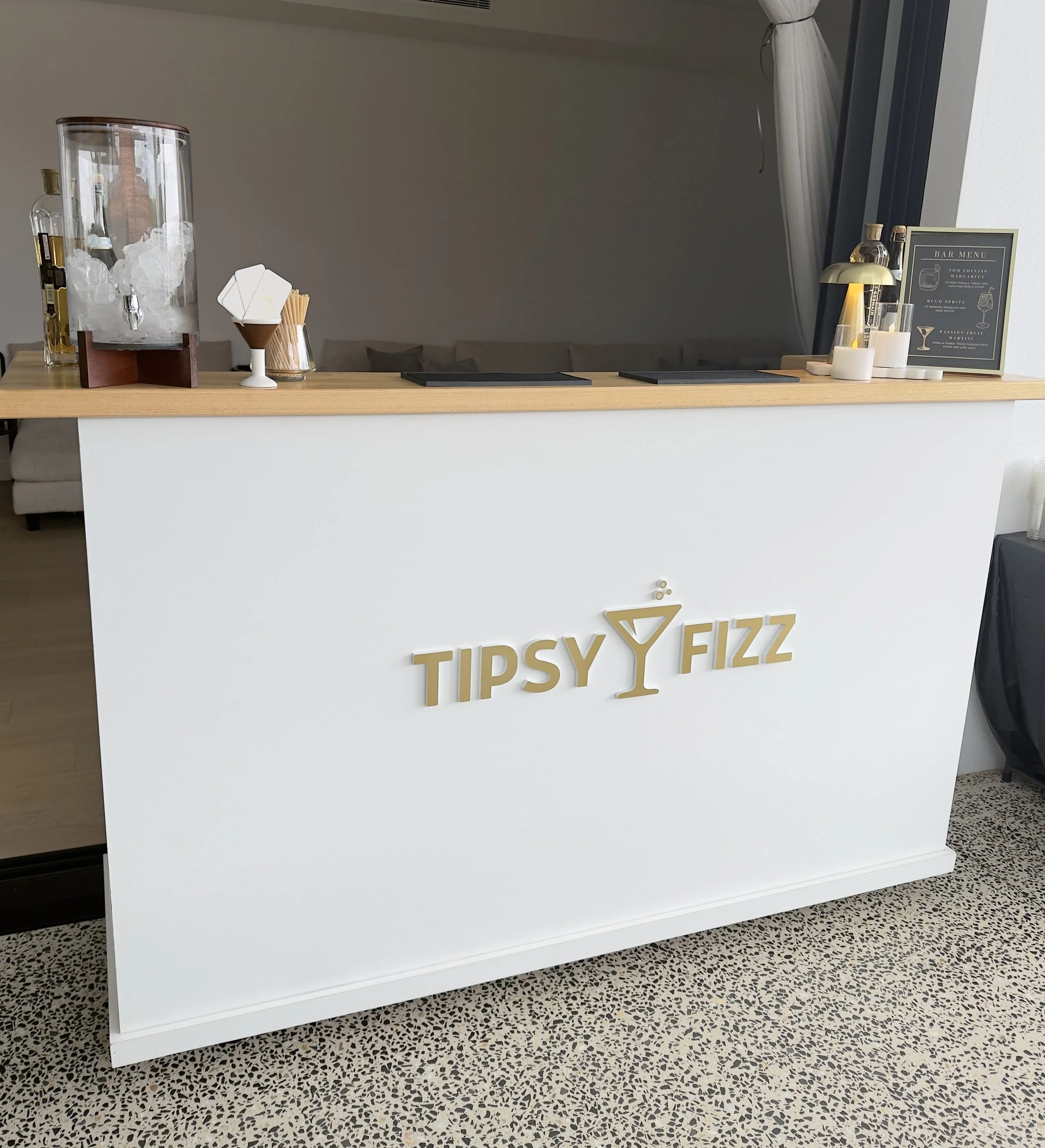 A white bar counter with the words 'TIPSY FIZZ' in gold letters. On the counter, there is a water dispenser filled with ice, a small white container with paper straws, a framed bar menu, and candles. The background includes a wall with a window and curtained opening.