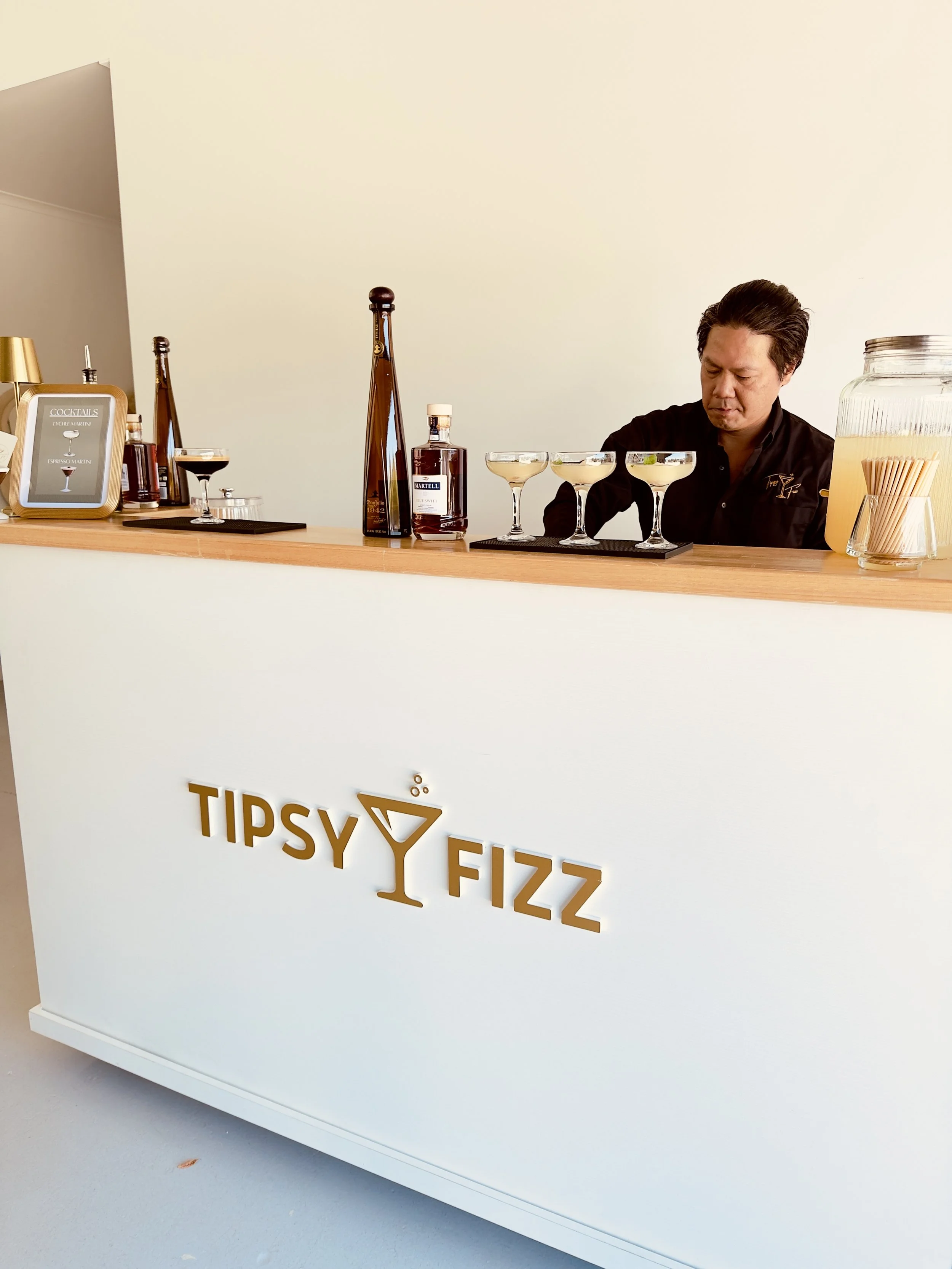 A bar counter with bottles of liquor, glasses with cocktails, and a man preparing drinks. Part of a sign with the words 'TIPSY Y FIZZ' is visible on the front of the counter.