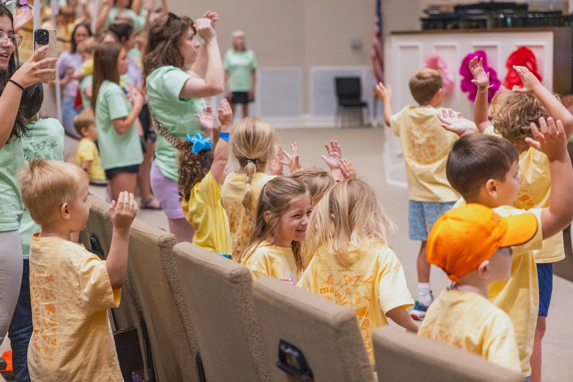 A group of children in yellow shirts, some sitting and some standing, raising their hands in the Worship Center, with women at the front leading a dance.