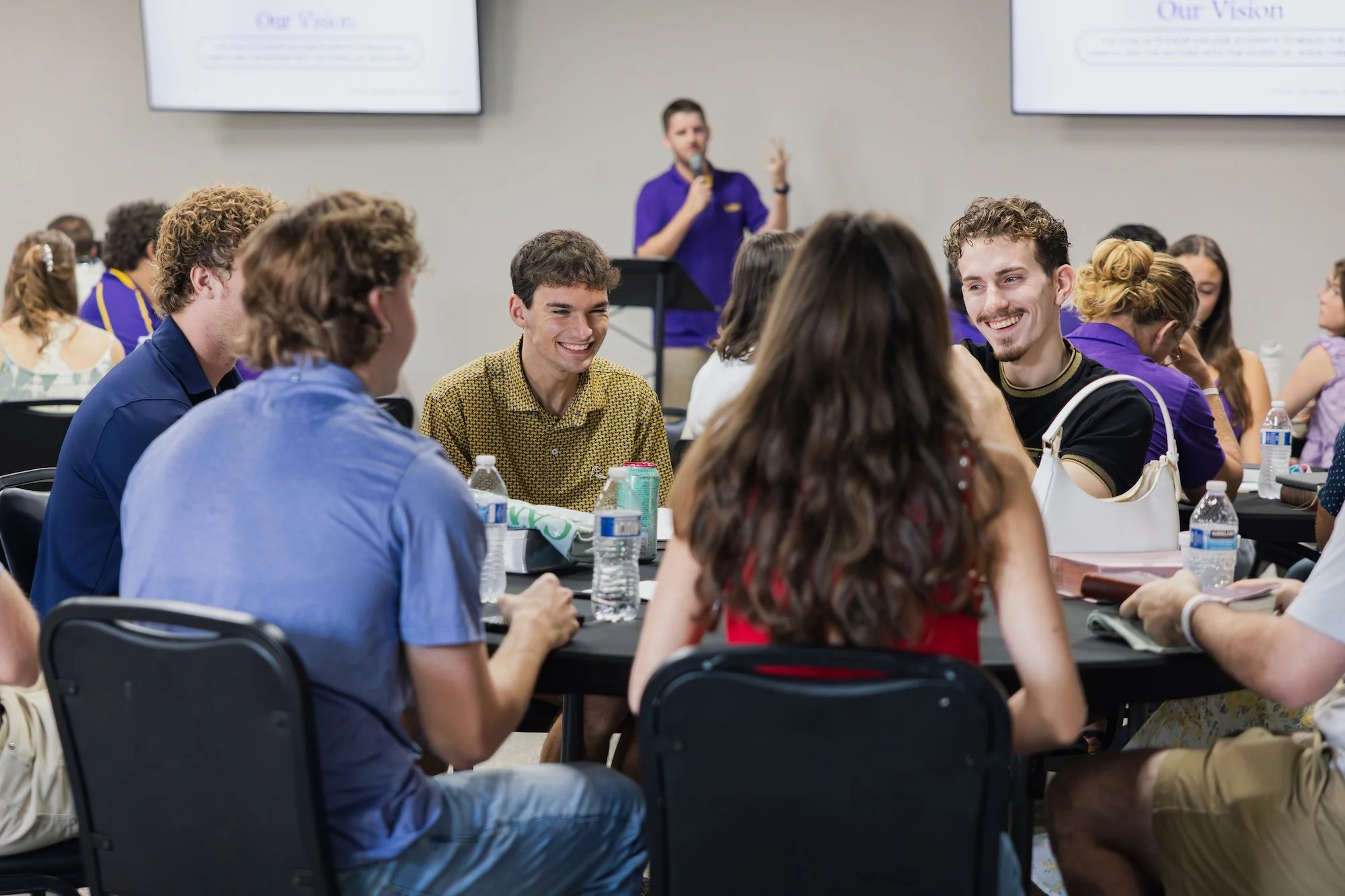 Group of people sitting around tables, smiling and engaging in conversation during a seminar or conference, with a presenter speaking at a podium in the background.