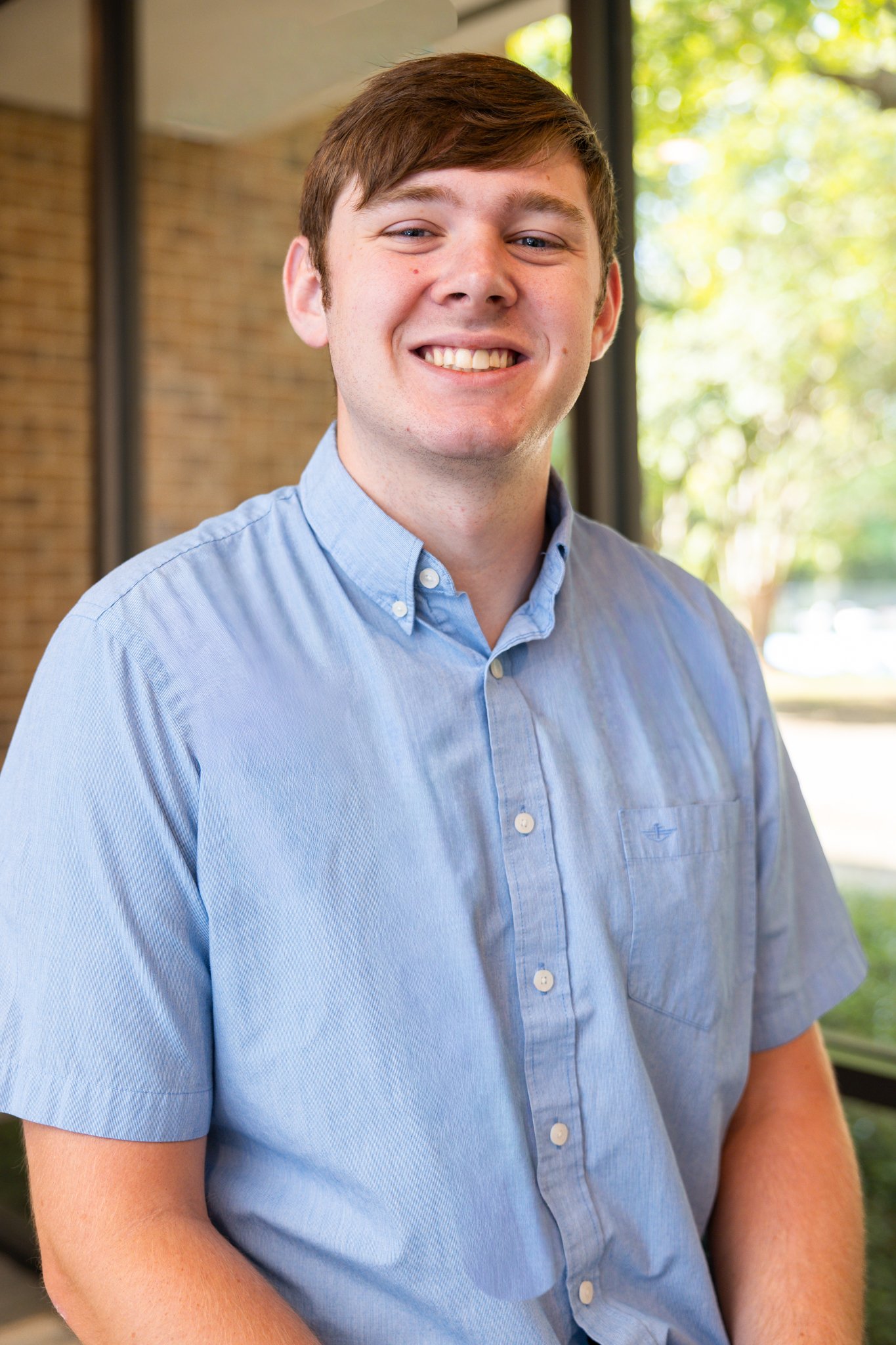 A young man with short brown hair smiling and wearing a beige polo shirt with a small logo, sitting in front of a blue background.