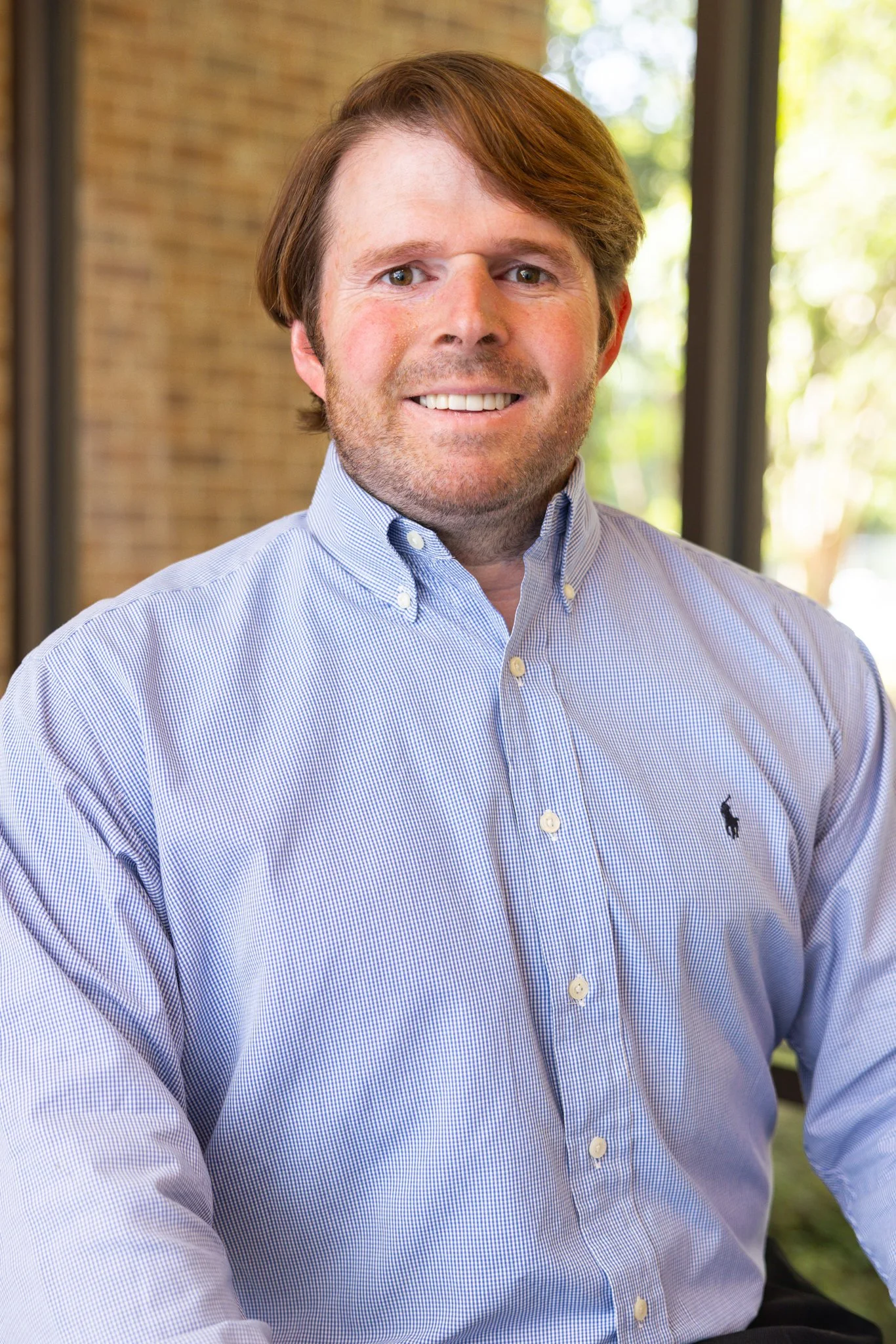 A smiling man with brown hair and a beard wearing a light blue button-up shirt standing in front of a blue background with blurred seats.