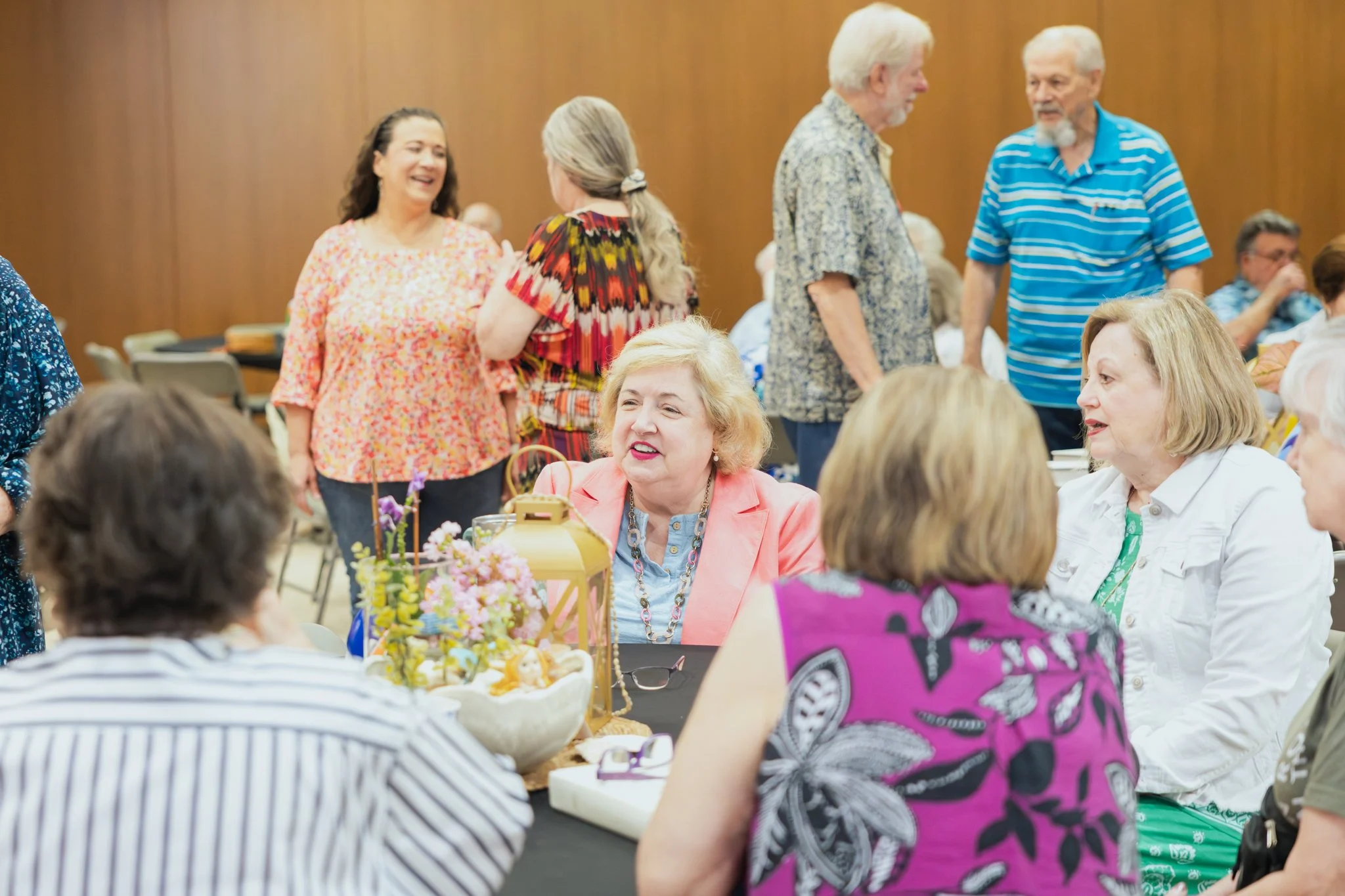Group of elderly people socializing at a gathering, some women seated at a table decorated with flowers and food, others standing and talking in a room with wooden walls.