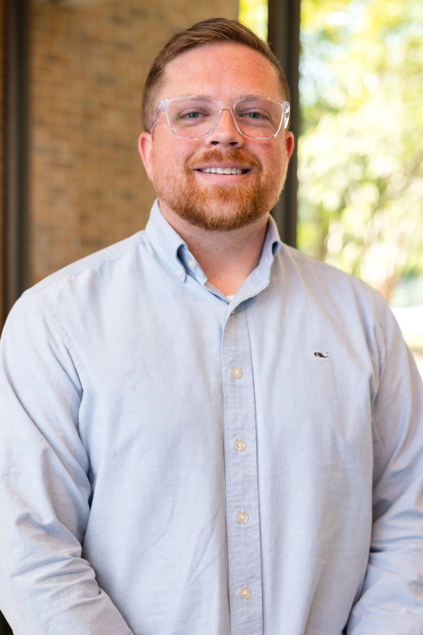 Portrait of a smiling man wearing glasses and a light-colored button-up shirt, standing in front of a blurred background of blue seating.