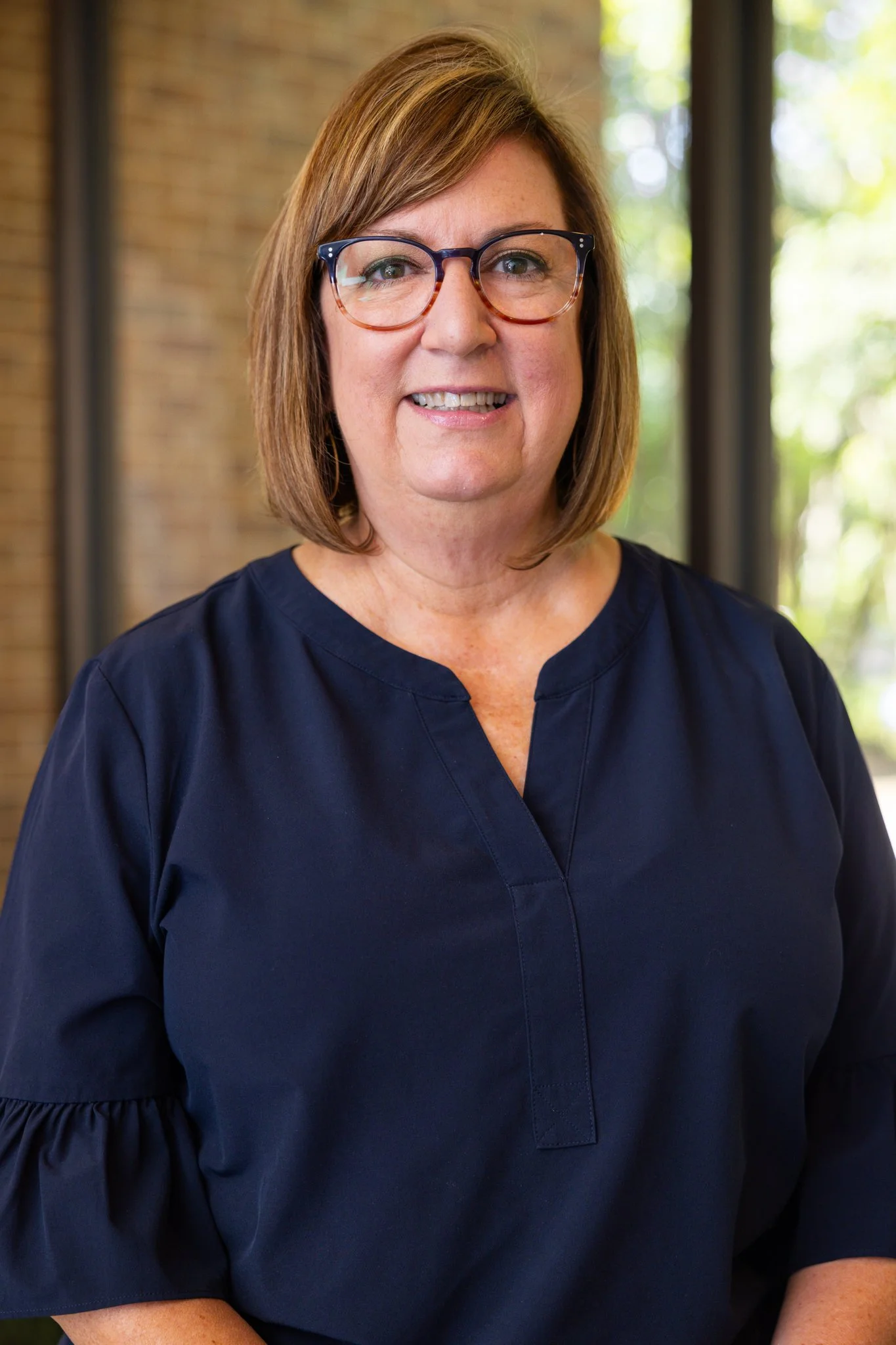 A woman with shoulder-length light brown hair, wearing glasses, smiling, and dressed in a white and navy striped blouse, standing against a blurred blue background.