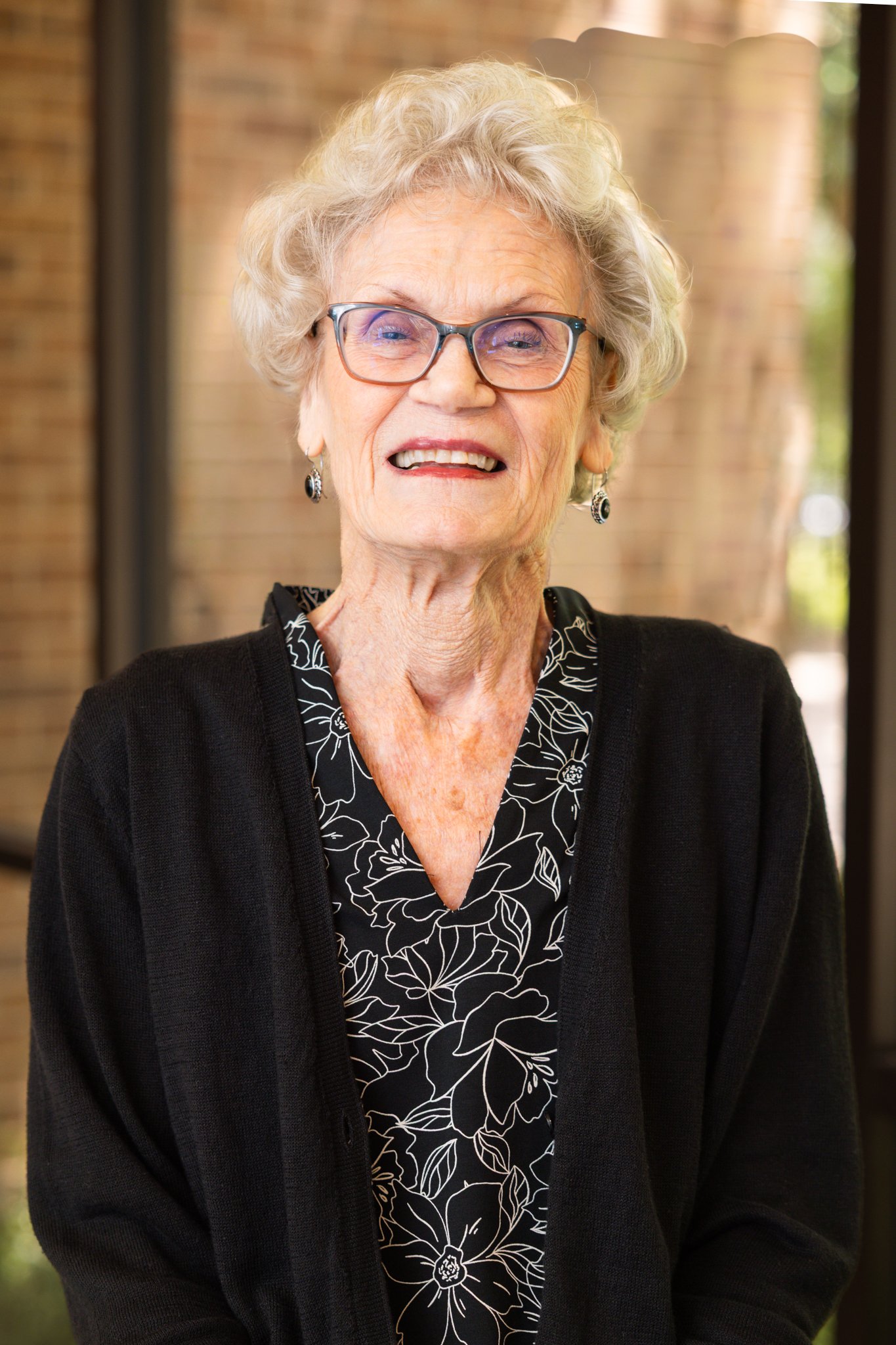An elderly woman with gray hair styled in a short, wavy manner, smiling and wearing a gray blazer and white top, against a blurred blue background.