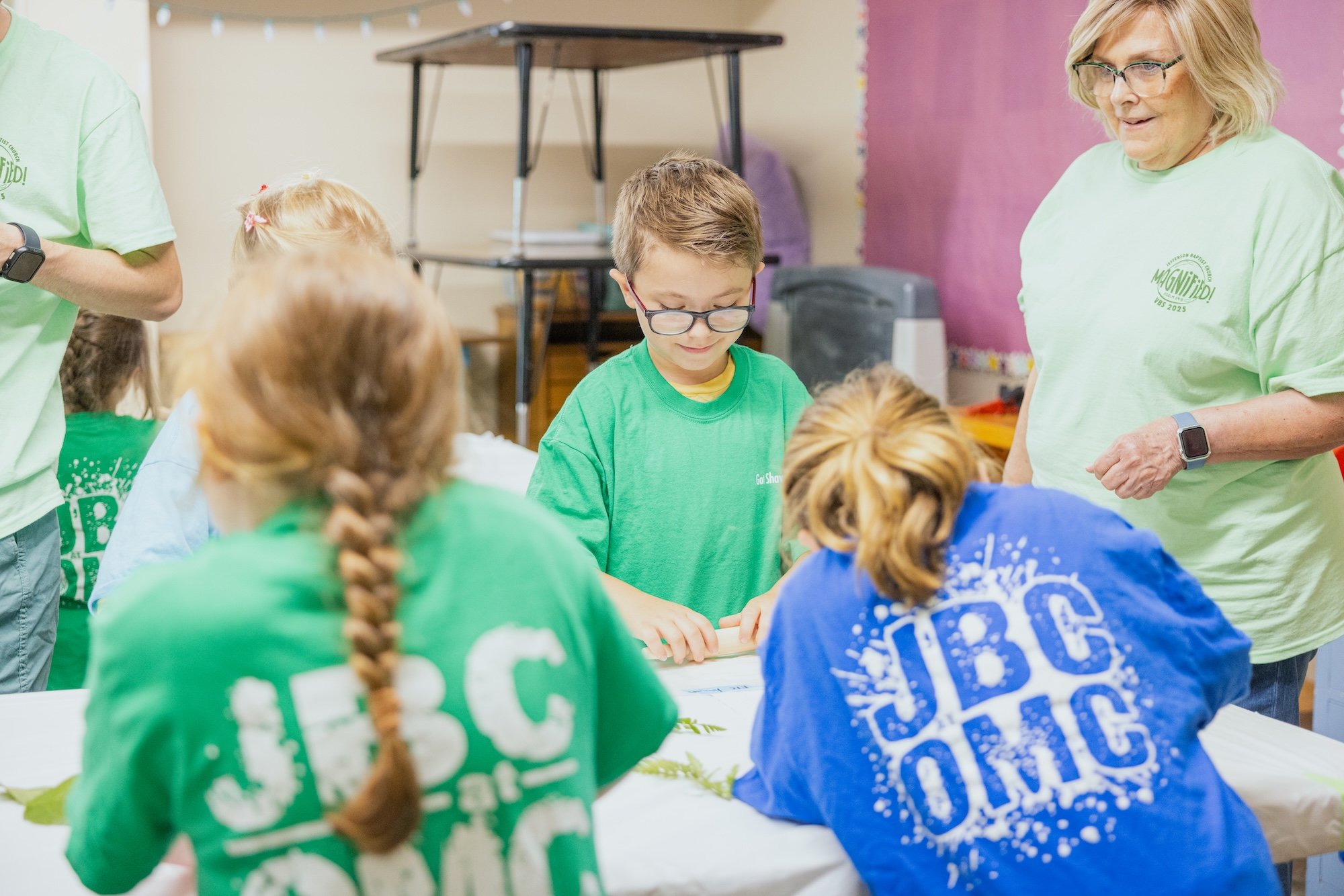 Children sitting at a table participating in a craft activity, with an adult supervising in a classroom setting.