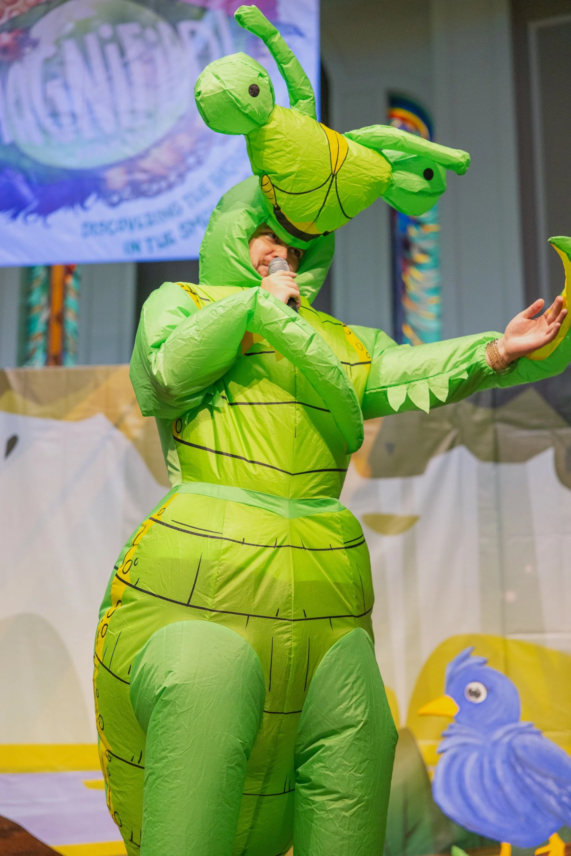 A person dressed in a large, bright green inflatable praying mantis costume, holding a microphone and speaking on stage. Behind them is a backdrop with colorful cartoonish decorations.