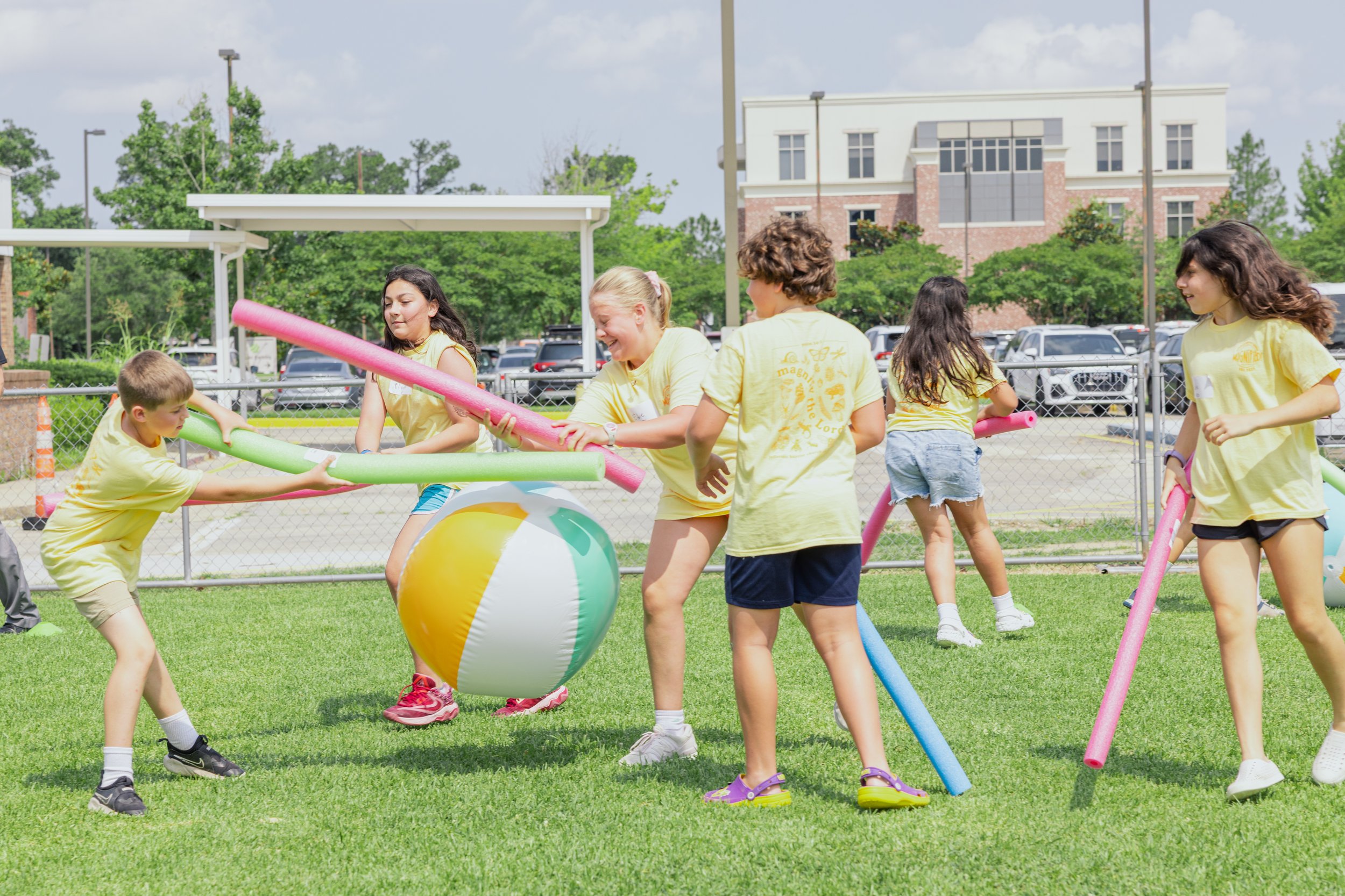Children are playing with pool noodles and a beach ball outdoors on a grassy field, with a chain-link fence and trees in the background.