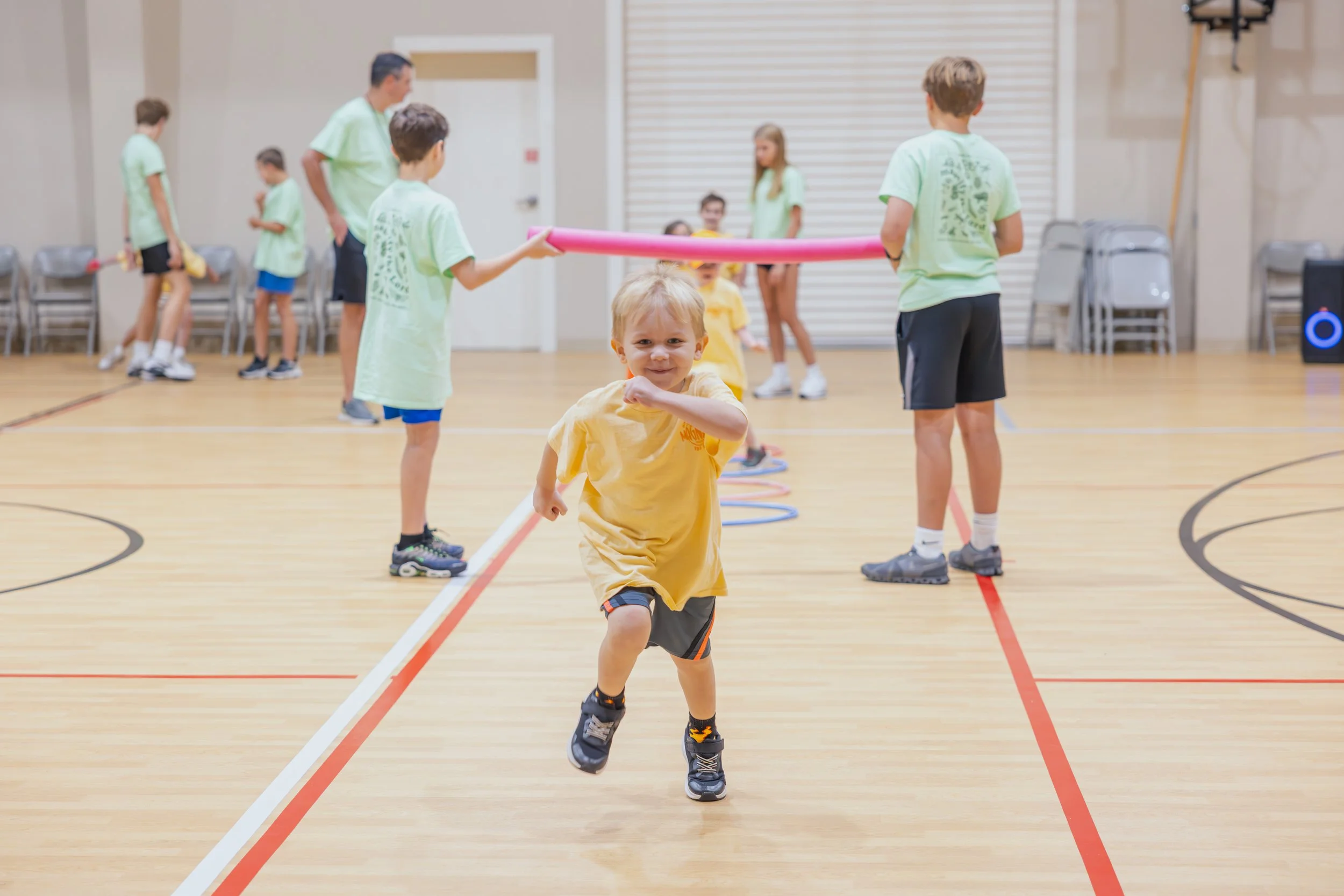 Young child running and smiling with a group of children and adults in a gymnasium, some holding foam noodles and participating in an activity.