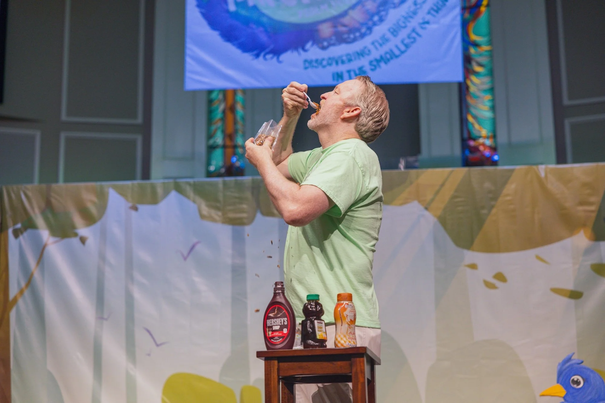 A man with gray hair, wearing a light green shirt, is standing at a table eating food with a spoon. On the table are bottles of Hershey's syrup, a bottle of chocolate syrup, a bottle of caramel syrup, and an orange and white can. Behind him, there is a decorative backdrop with colorful patterns and a blue cartoon character with a yellow beak in the corner.