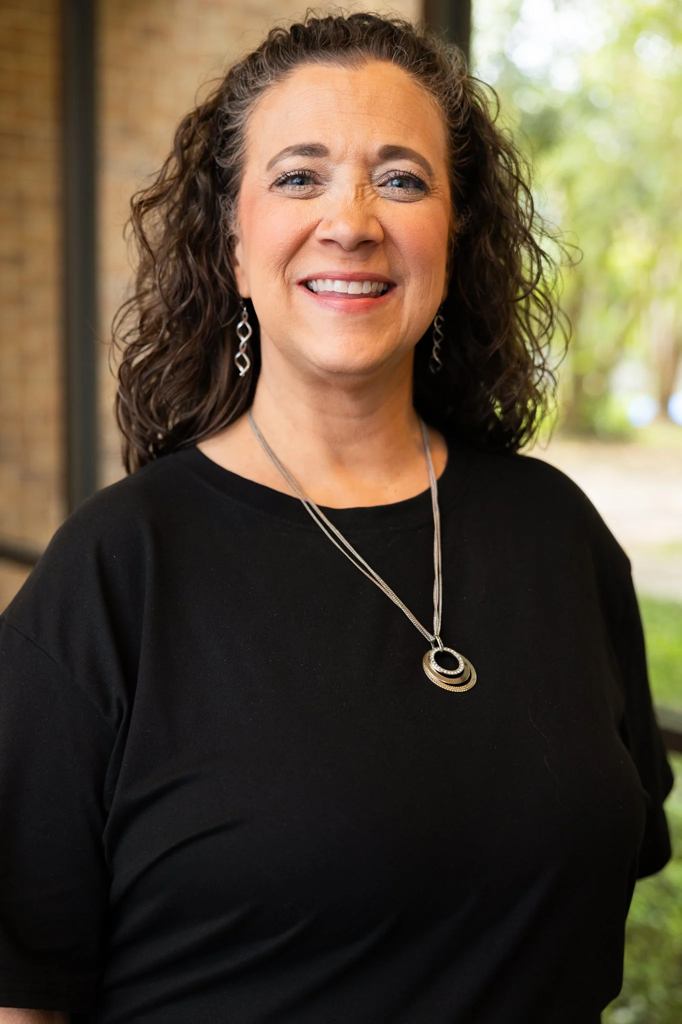 A smiling woman with curly dark hair wearing a black and white patterned jacket and jewelry stands against a blue background with dark columns.