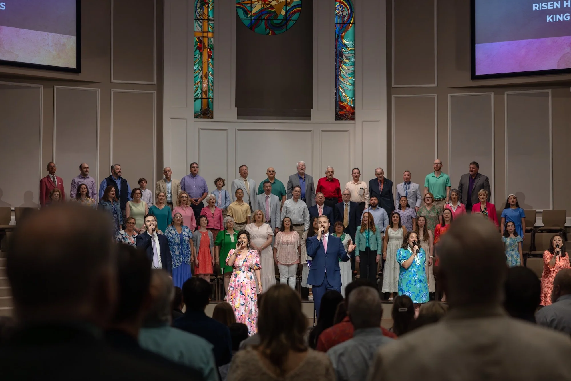 A congregation inside a church during a religious service, with singers and a choir on stage, stained glass windows behind them, and a TV screen displaying song lyrics.