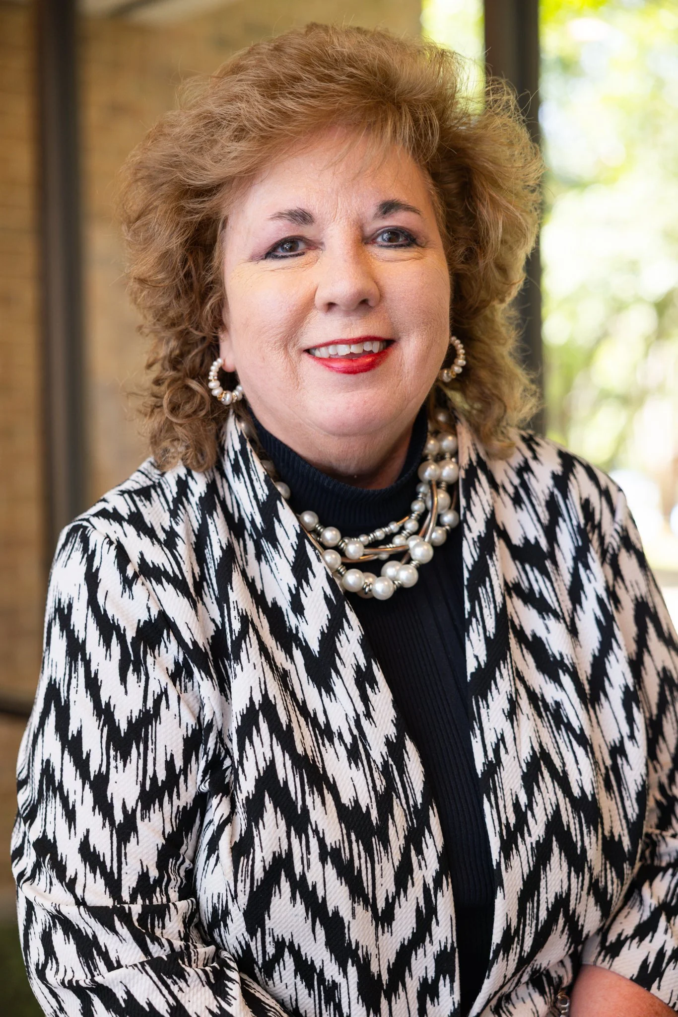 A woman with curly brown hair smiling, wearing a black and white striped jacket, black top, turquoise earrings, and a decorative necklace, standing in front of a blue abstract background.
