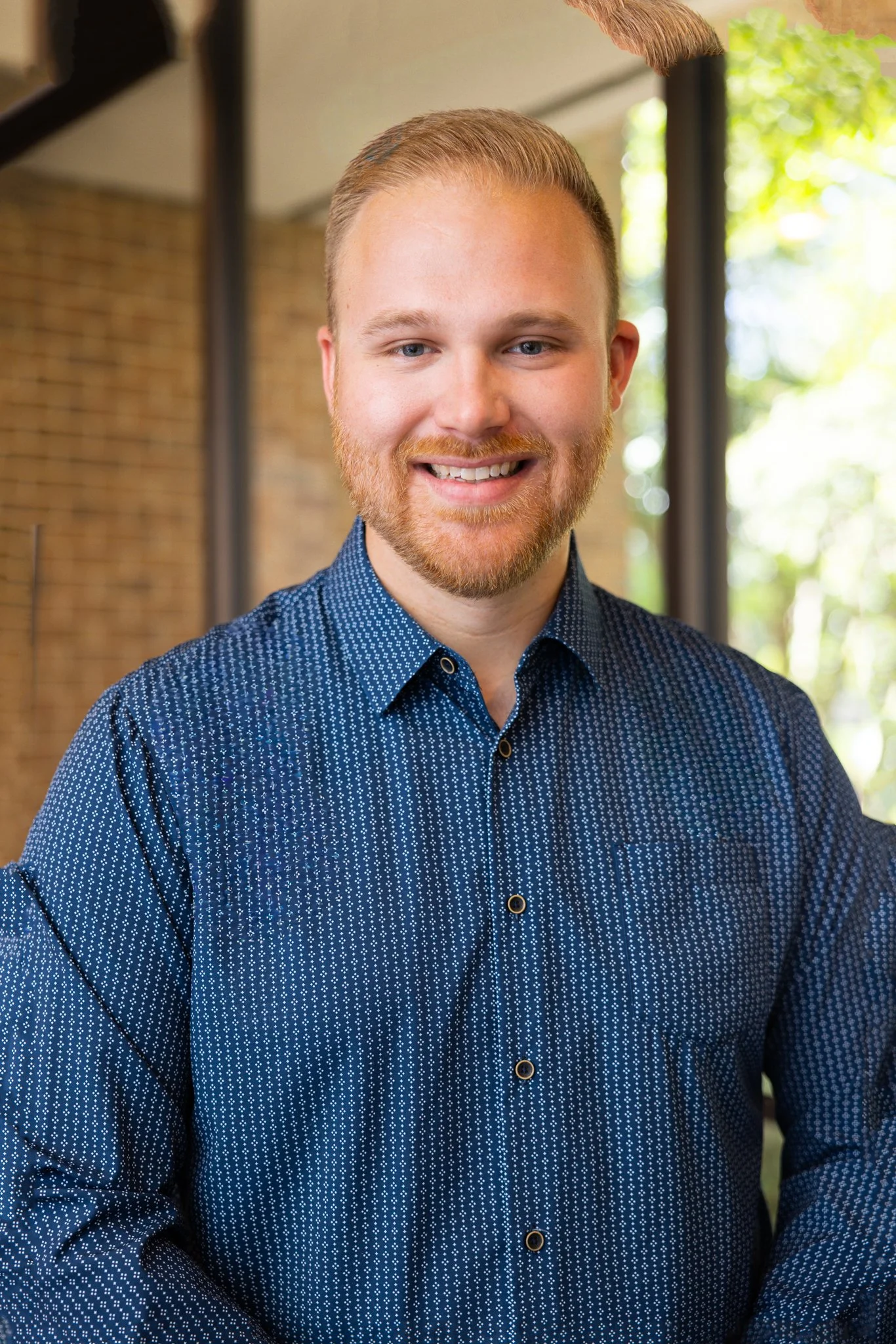 Headshot of a young man with blonde hair and beard, smiling, wearing a dark, patterned shirt, in front of blue theater seats.